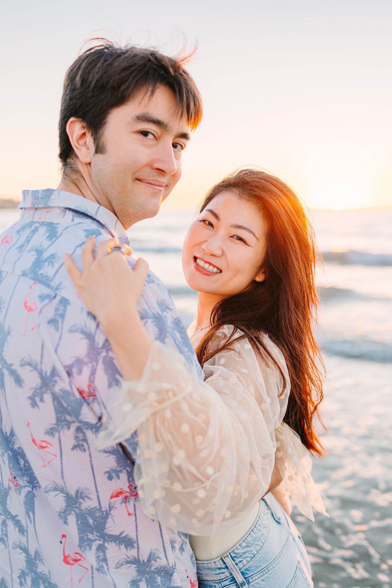 Happy couple in casual beachwear share an intimate moment while posing against ocean sunset backdrop.