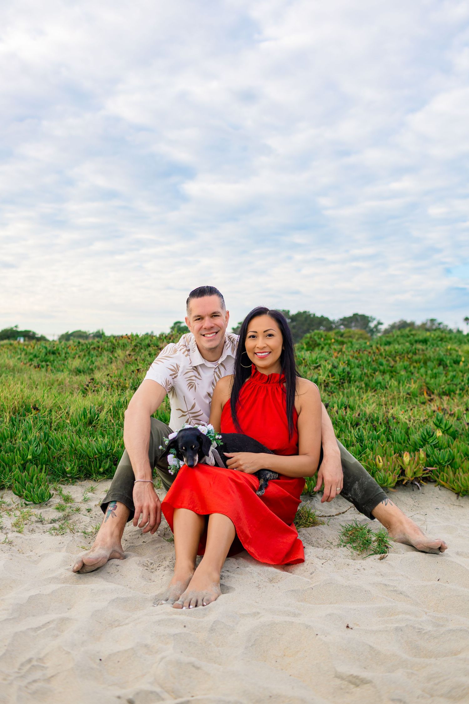 A couple relaxes together on a sandy beach with green foliage in the background during a sunny day.