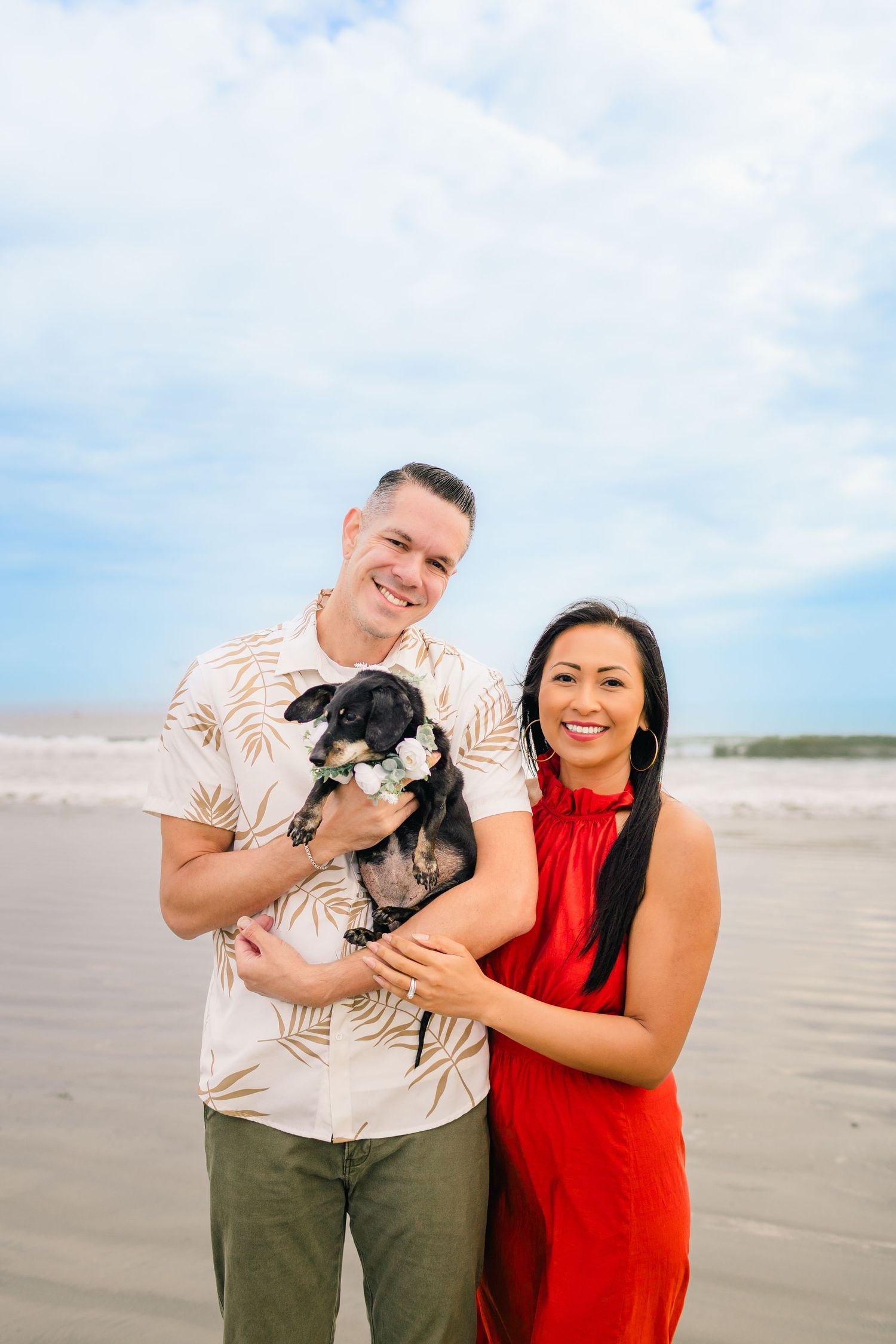 A couple in coordinated beachwear poses with their small black dog against a cloudy coastal backdrop.