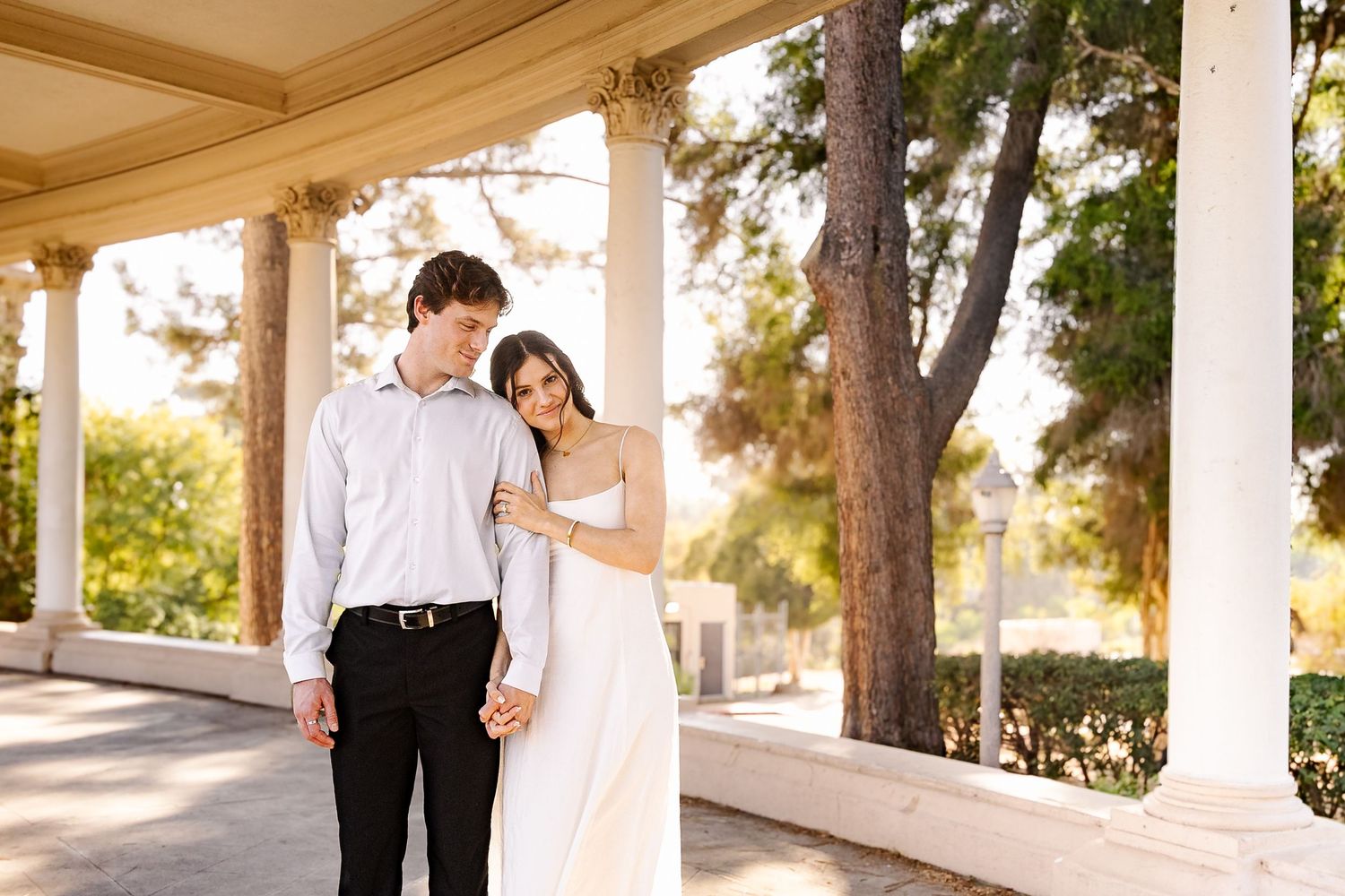 A couple in wedding attire walks along a covered colonnade with white pillars and trees in the background.