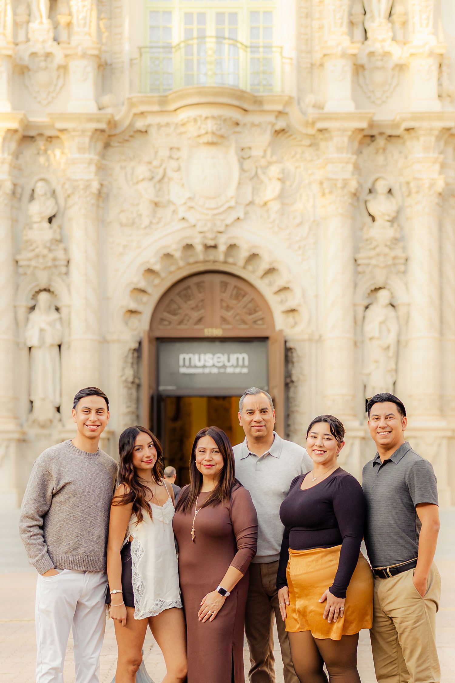 A group of people in coordinated earth-toned outfits pose together against a white stone architectural backdrop.