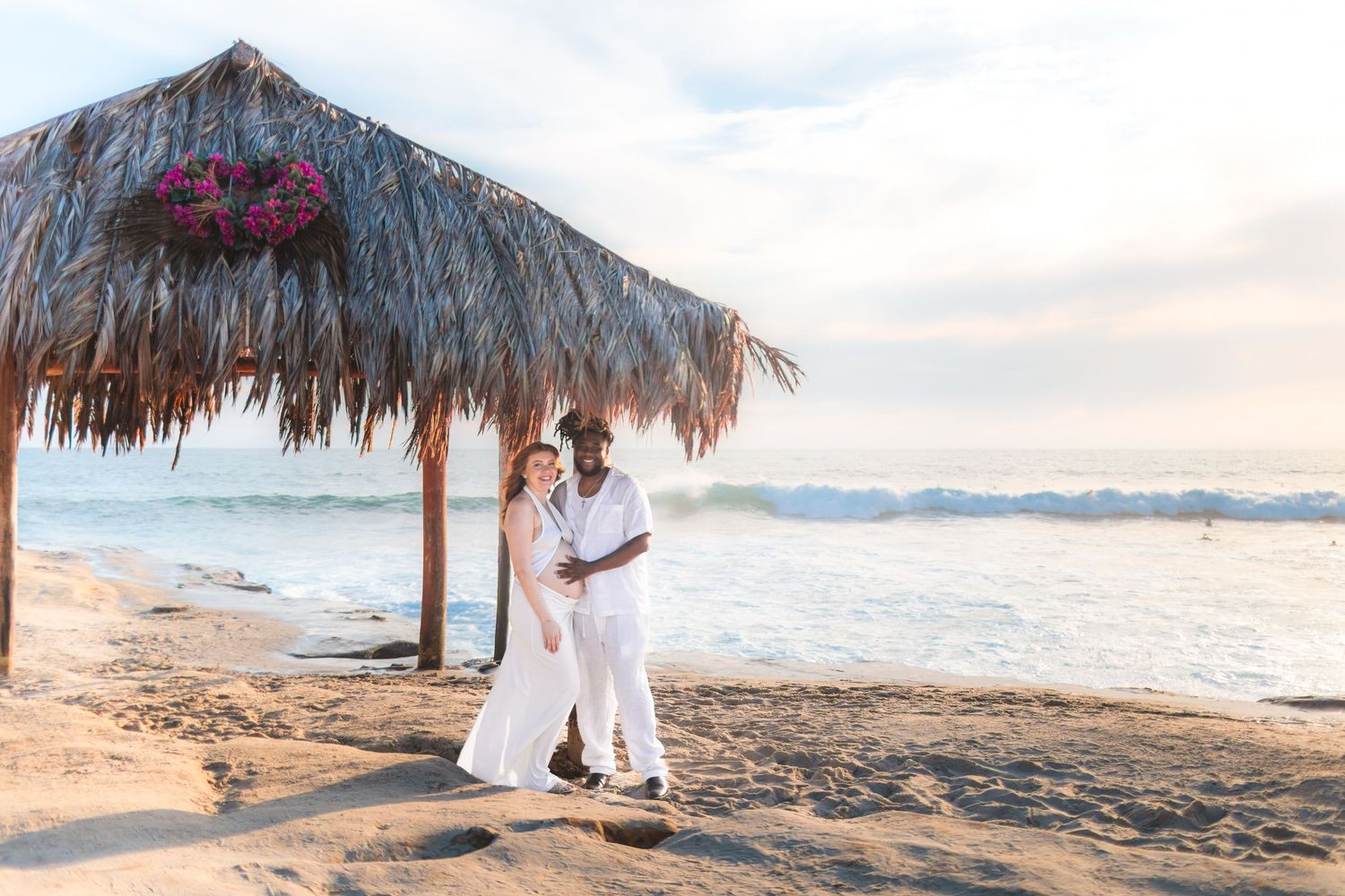 Couple in white outfits embracing on a scenic beach with thatched umbrellas at sunset.