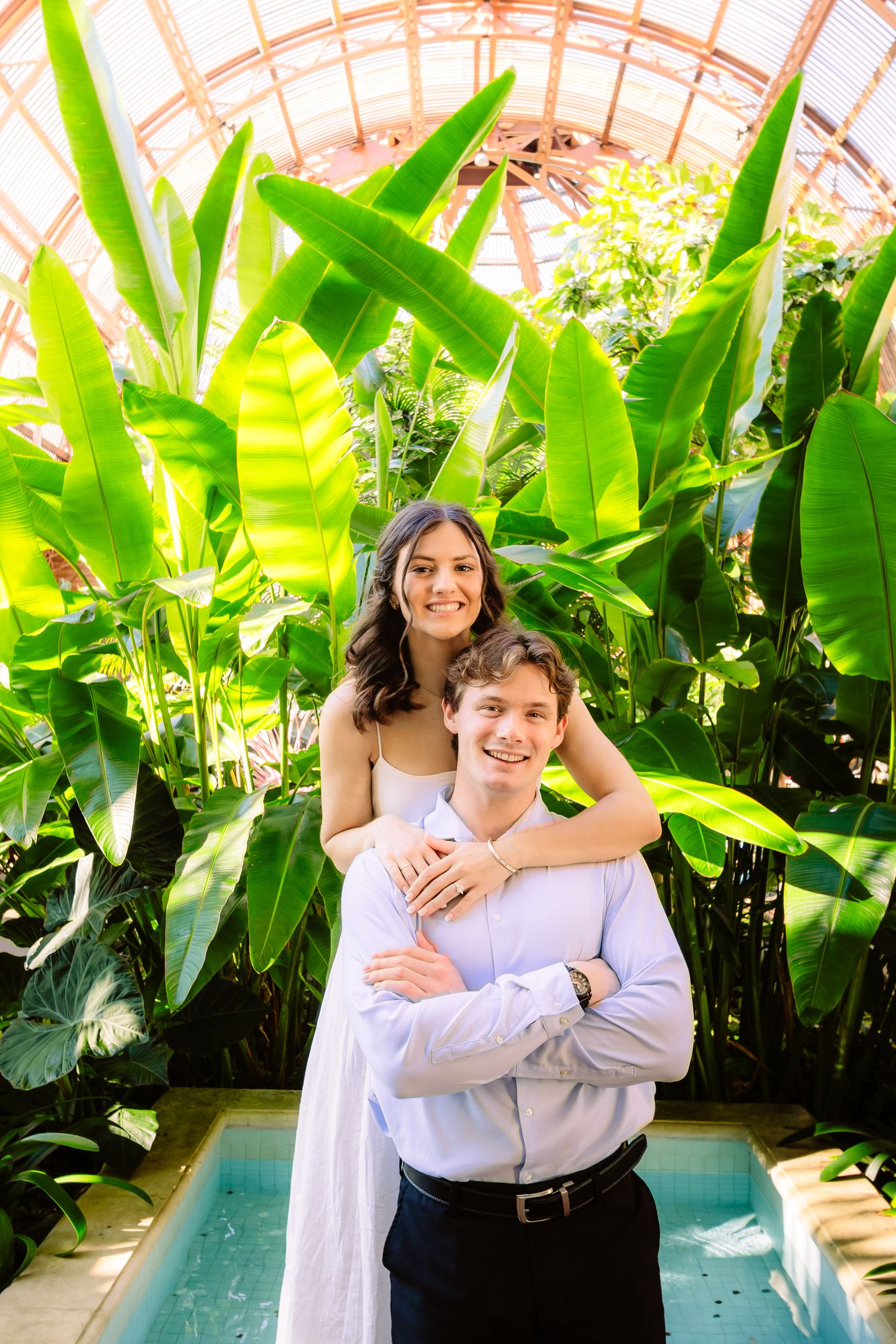 A couple embraces in front of tropical plants in a bright greenhouse setting.