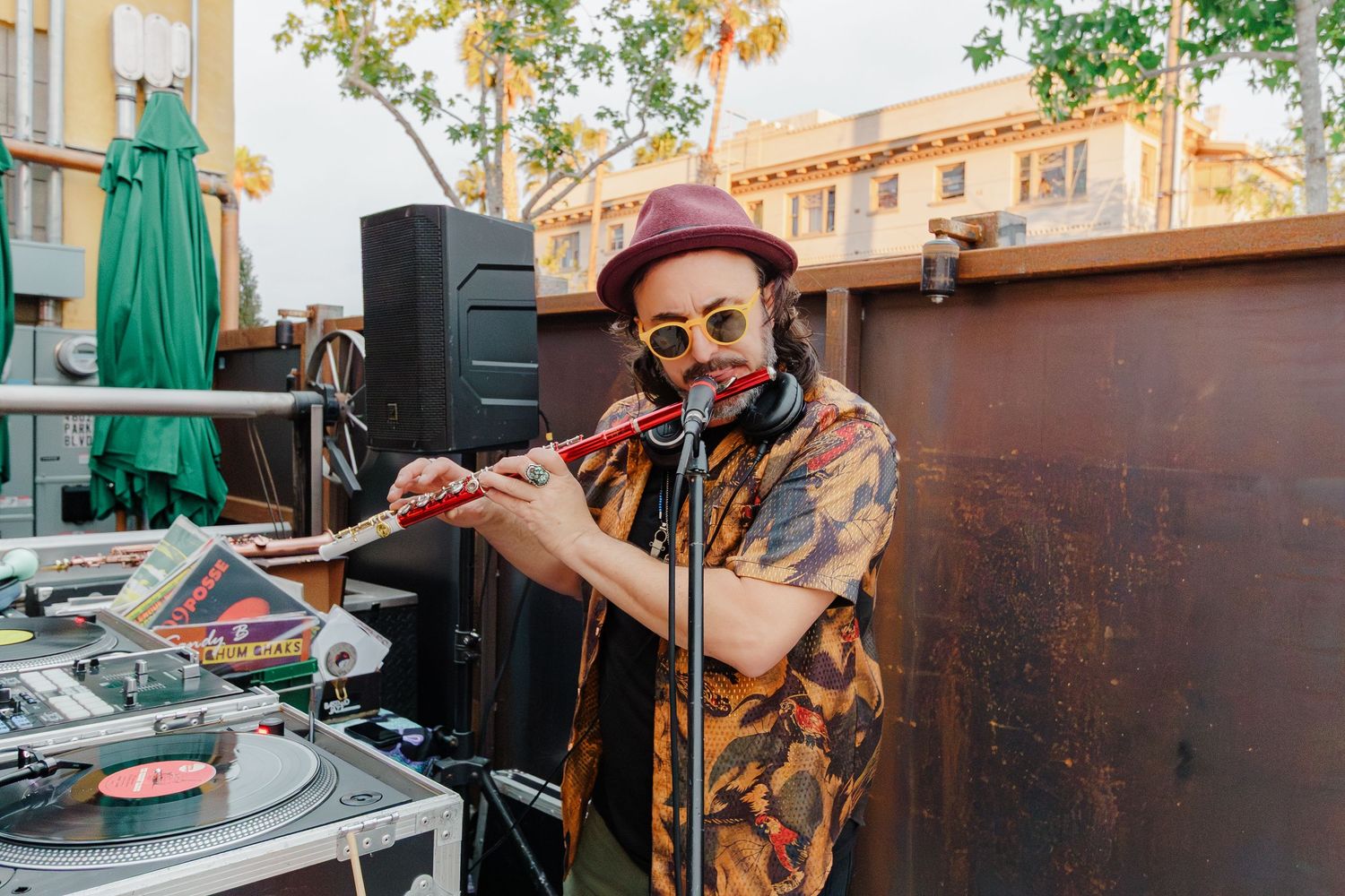 DJ performs with turntables at an outdoor venue with speakers and autumn trees in background.
