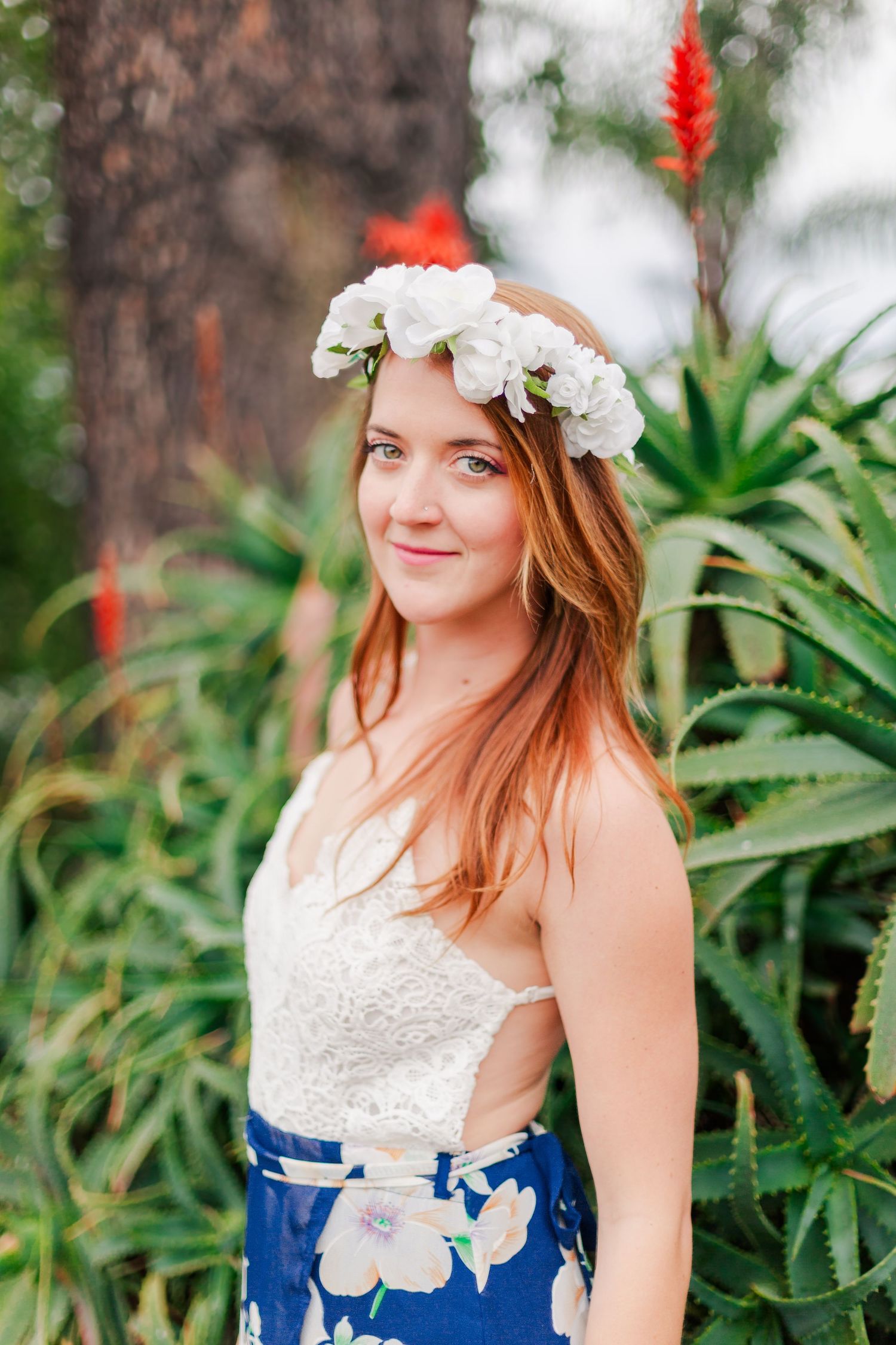 Woman wearing white floral crown and blue floral skirt poses in tropical garden setting.