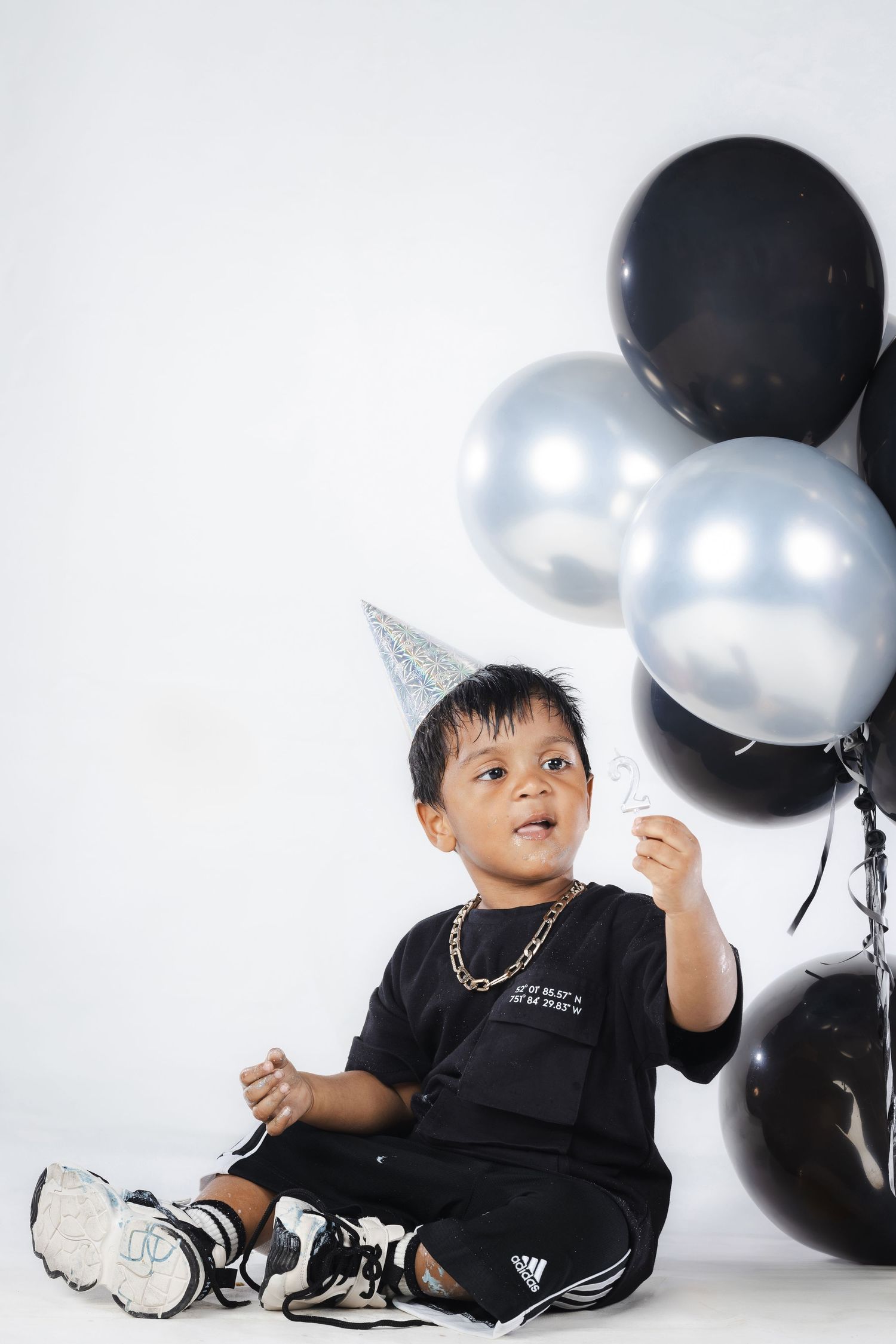 Child in party hat sitting with black and silver balloons against white background.