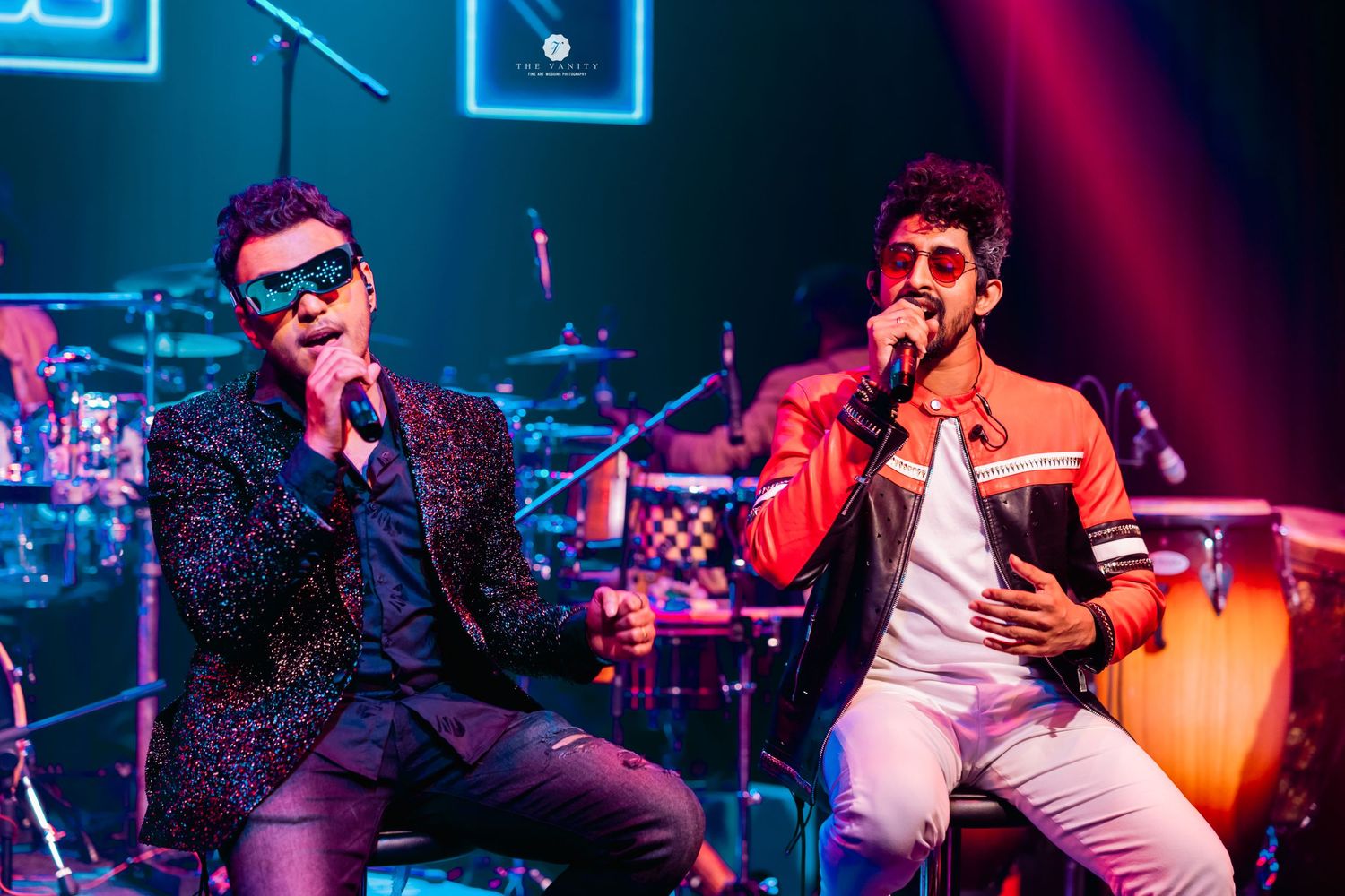 Two performers in flashy outfits singing on stage with neon lights and musical equipment in the background.