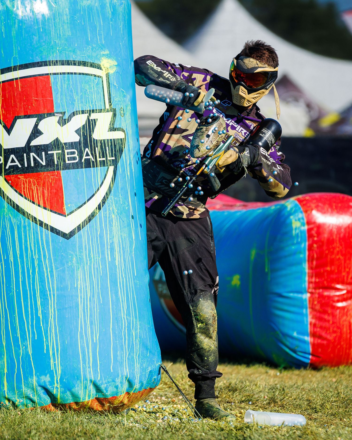 Player with paintball marker moves tactically past colorful NXL bunker during gameplay.