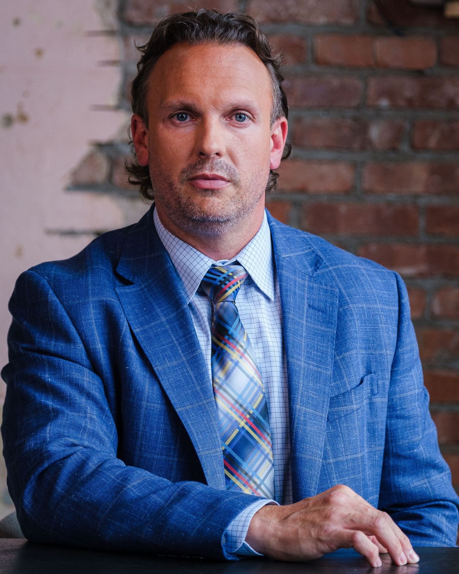 Professional portrait of person in blue plaid suit against brick wall background.