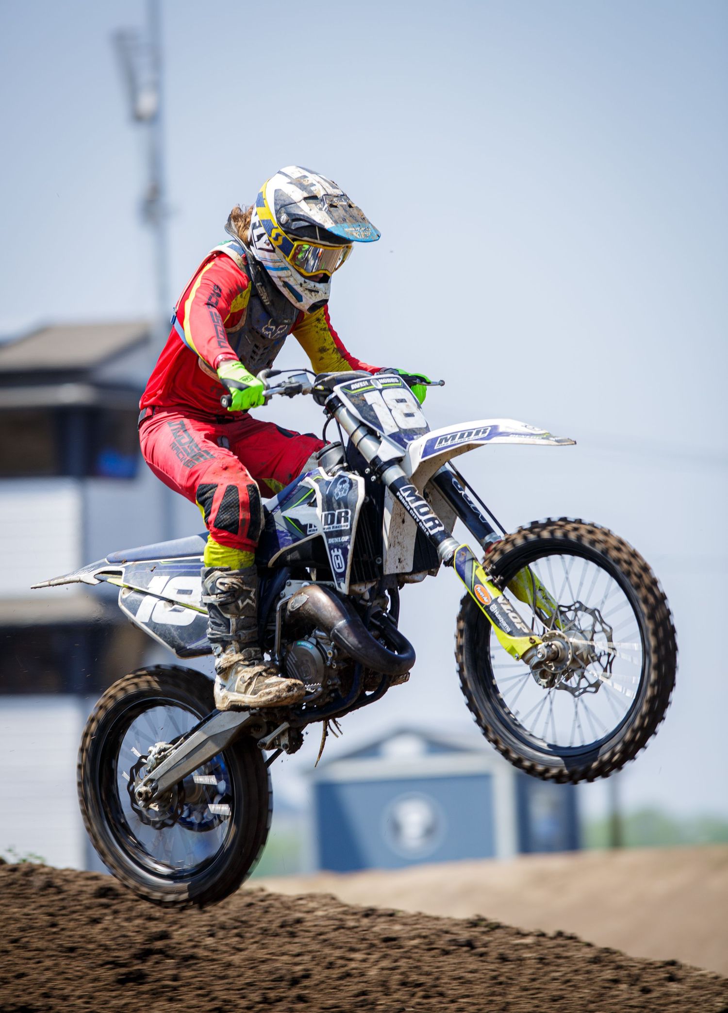 Dirt bike rider in red gear performs dramatic jump on motocross track against blue sky.