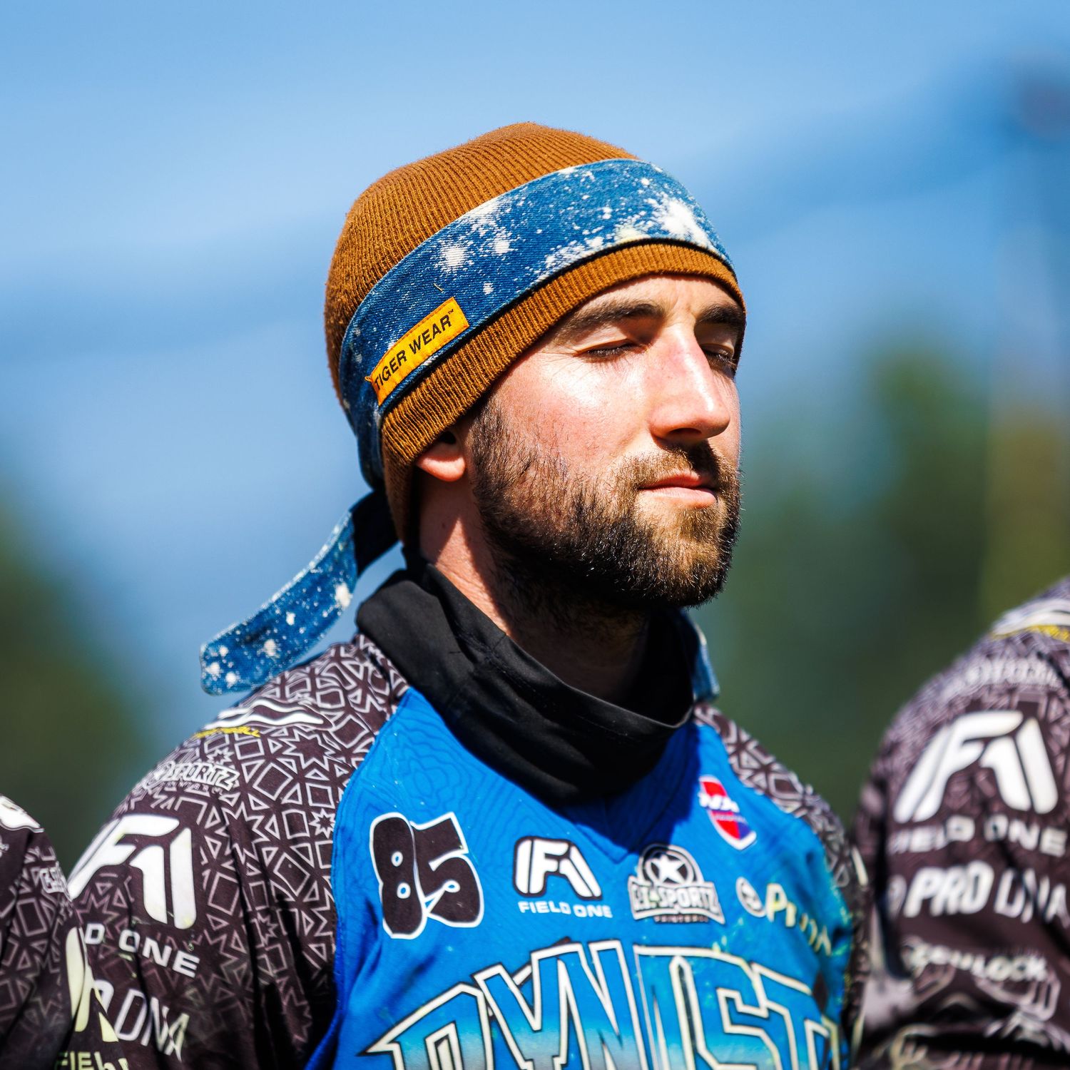 Paintball competitor wearing blue jersey and winter beanie during outdoor tournament.