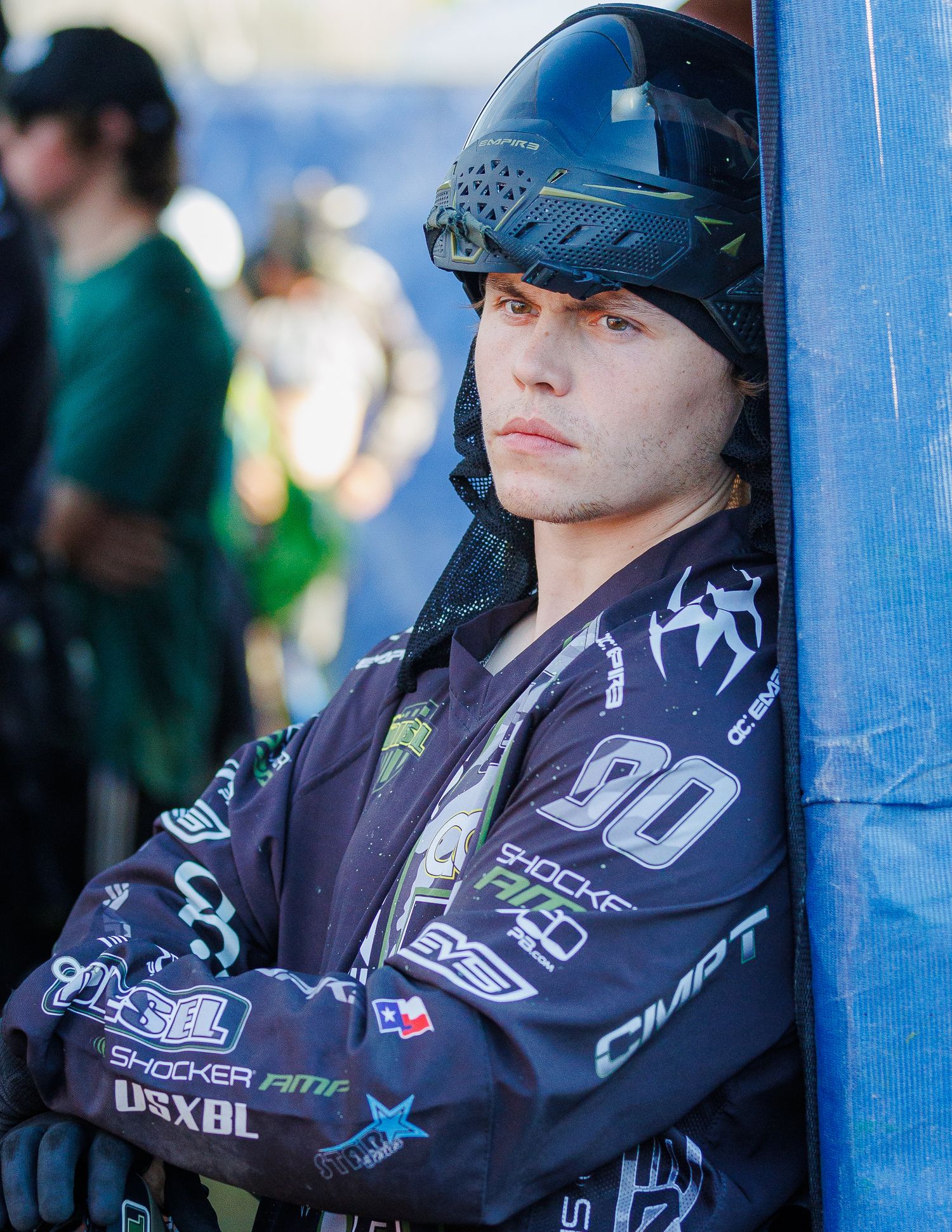 Paintball player wearing a blue helmet and black jersey with sponsors leans against a blue post.