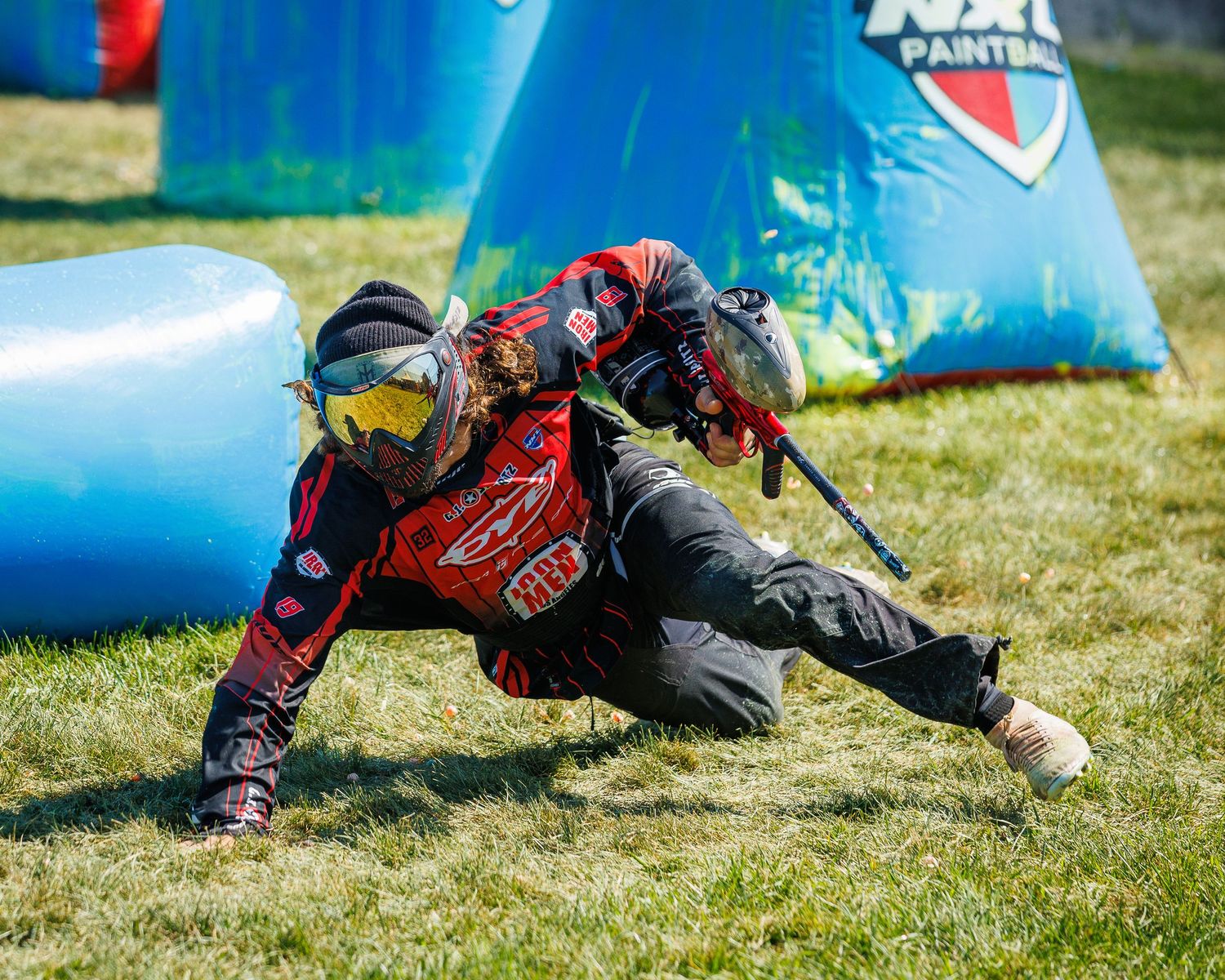 Paintball player in red jersey executing dynamic slide move behind blue bunker.