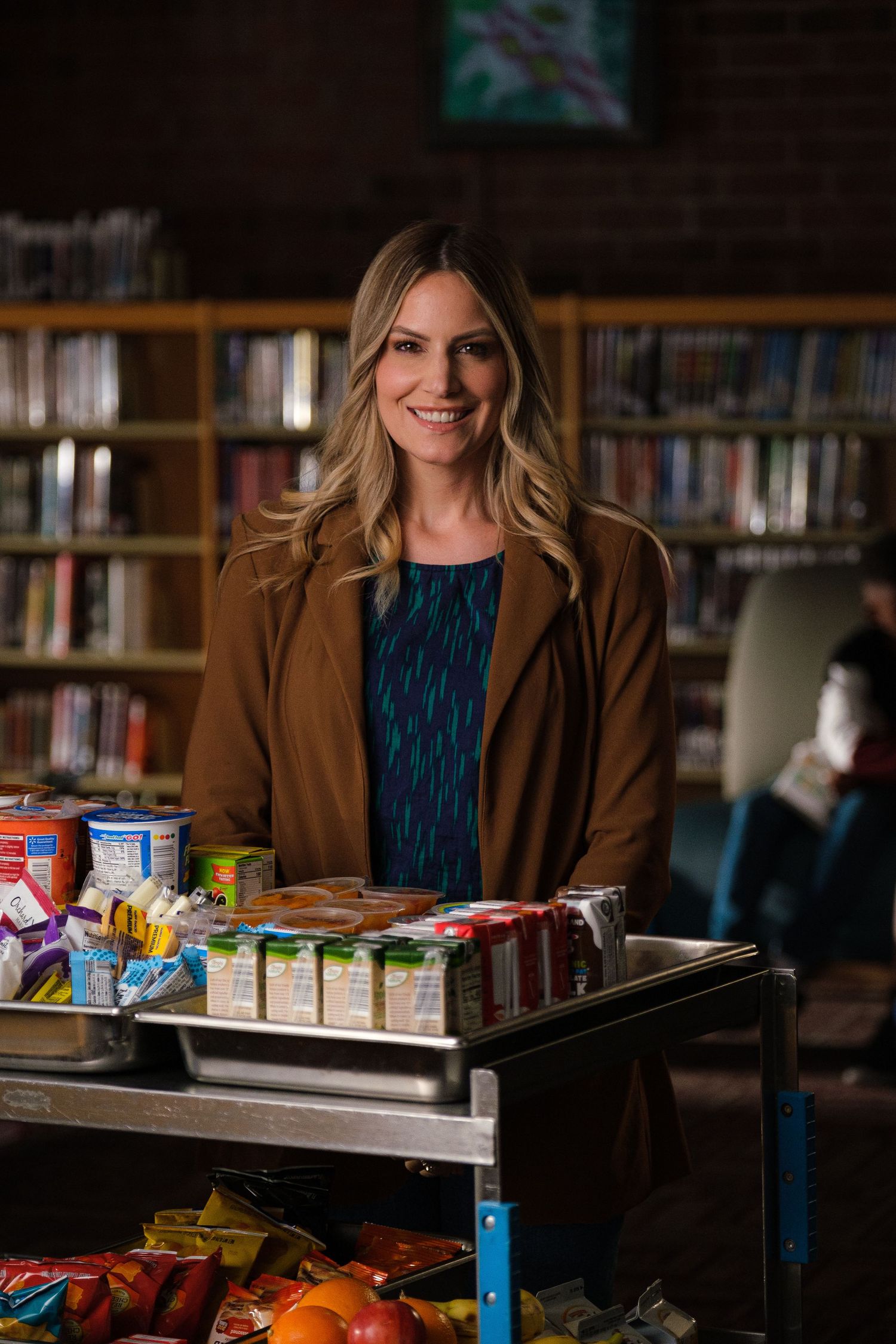 Food bank worker poses with cart of groceries and canned goods in library.