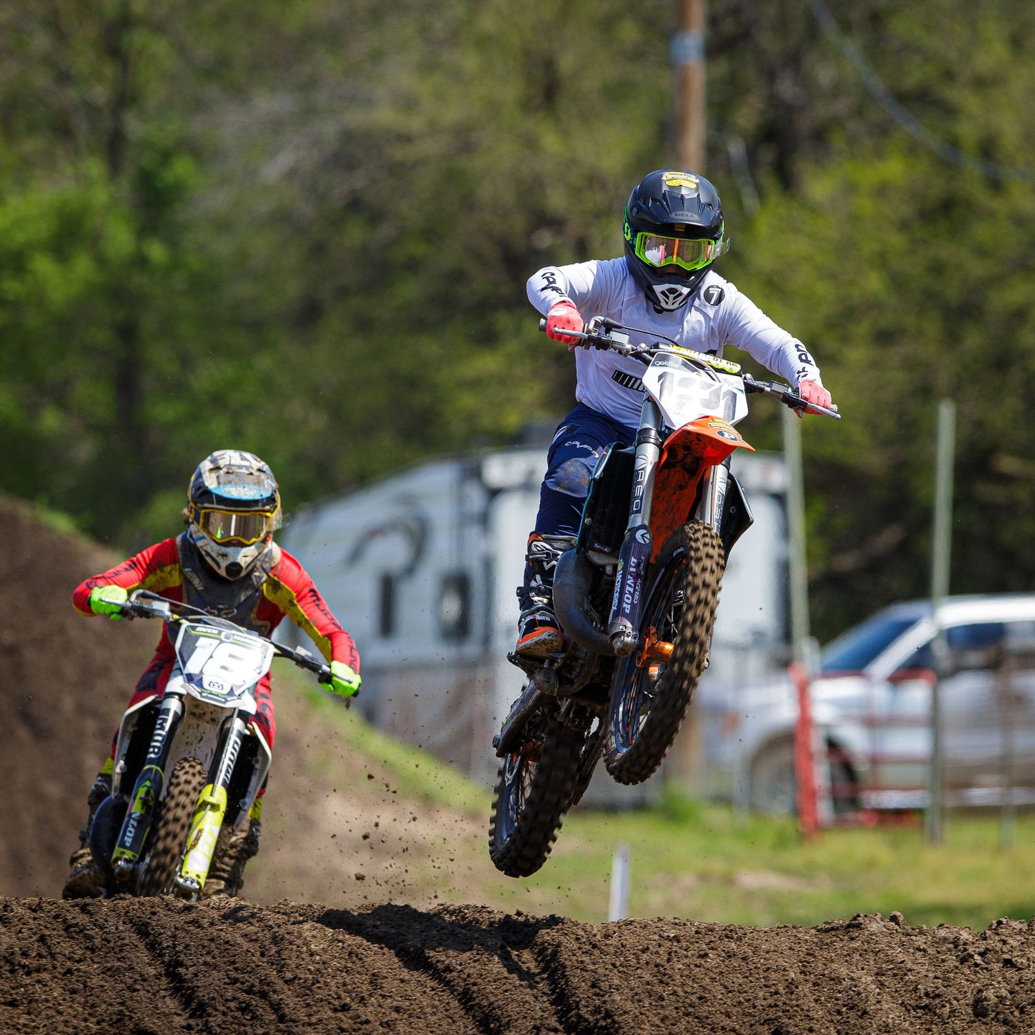 Two motocross riders jump their dirt bikes over a muddy track during an intense race competition.