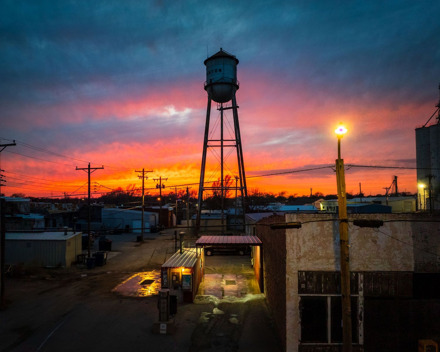 Vintage water tower silhouetted against vibrant sunset sky with street lights below.