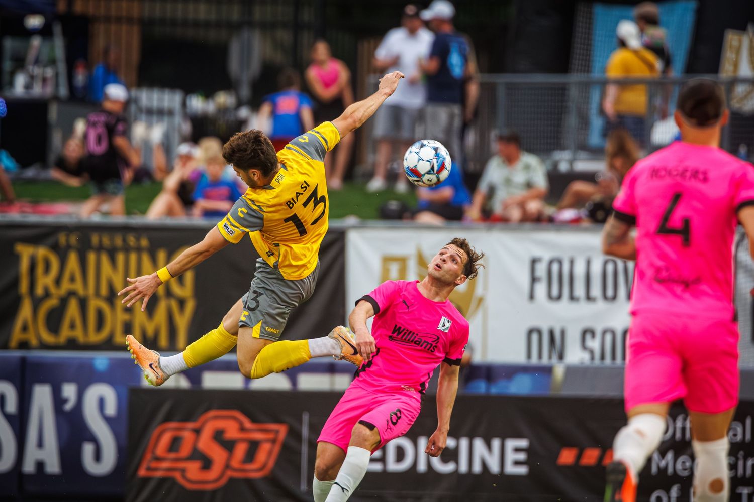Two soccer players leap into the air competing for the ball during a match between yellow and pink uniformed teams.