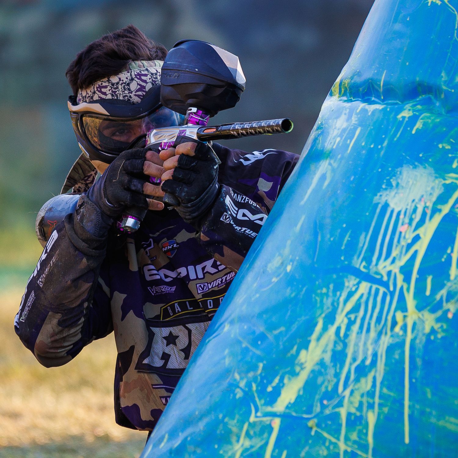 Paintball player takes aim from behind a blue inflatable bunker during competitive match.