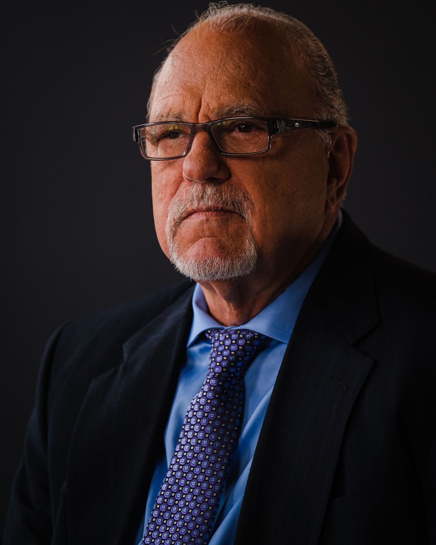 Corporate headshot of senior executive in dark suit and glasses against black background.