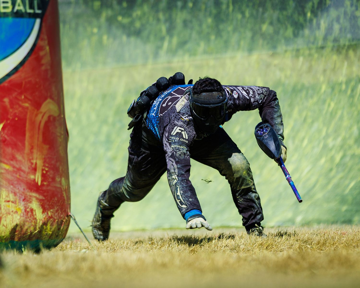 Player in black tactical gear slides low behind paintball bunker during intense match.