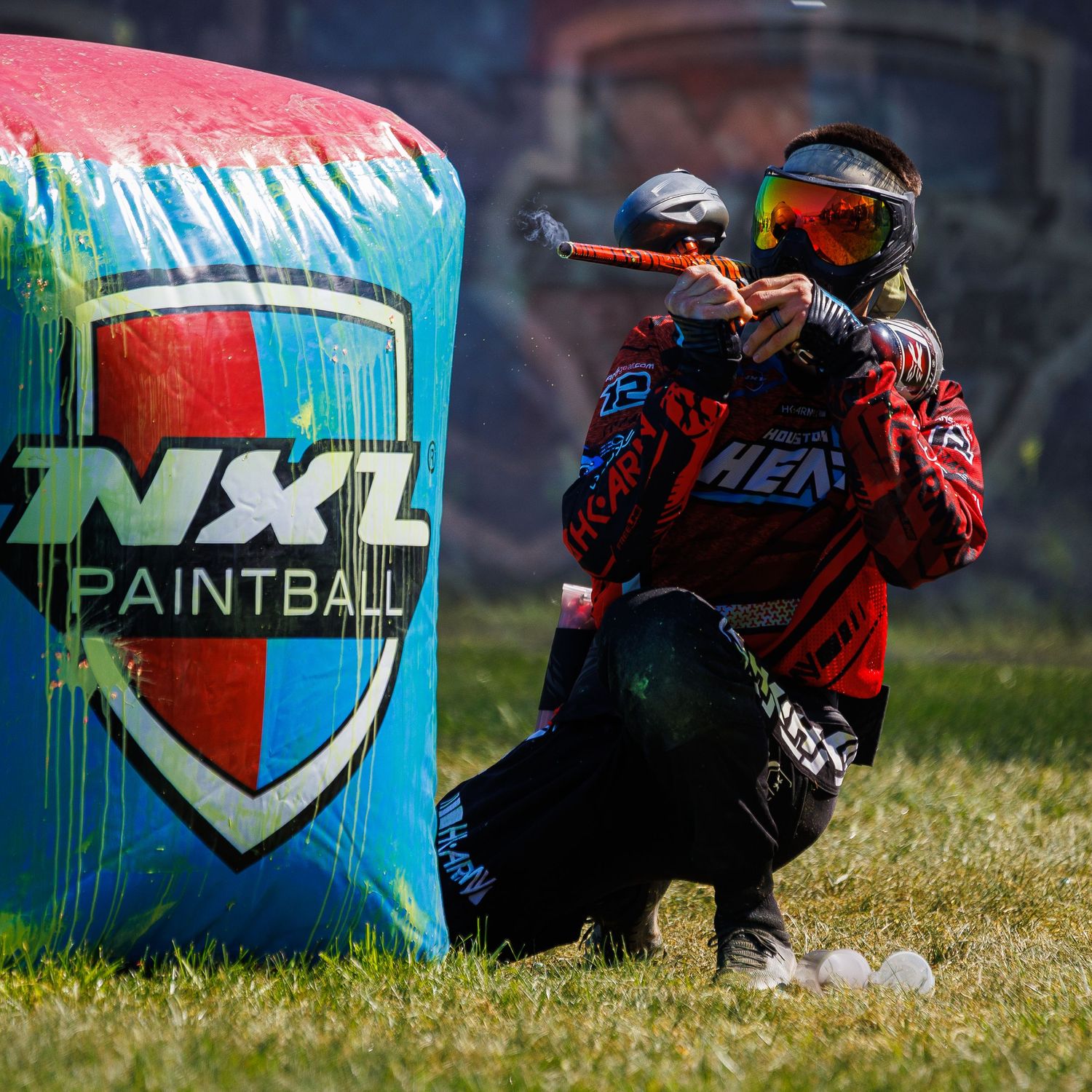 Paintball player crouches behind protective bunker with NXL logo during tournament play.