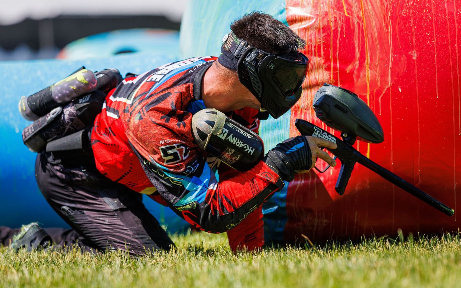 Paintball player crouching behind red bunker while aiming marker during competitive match.