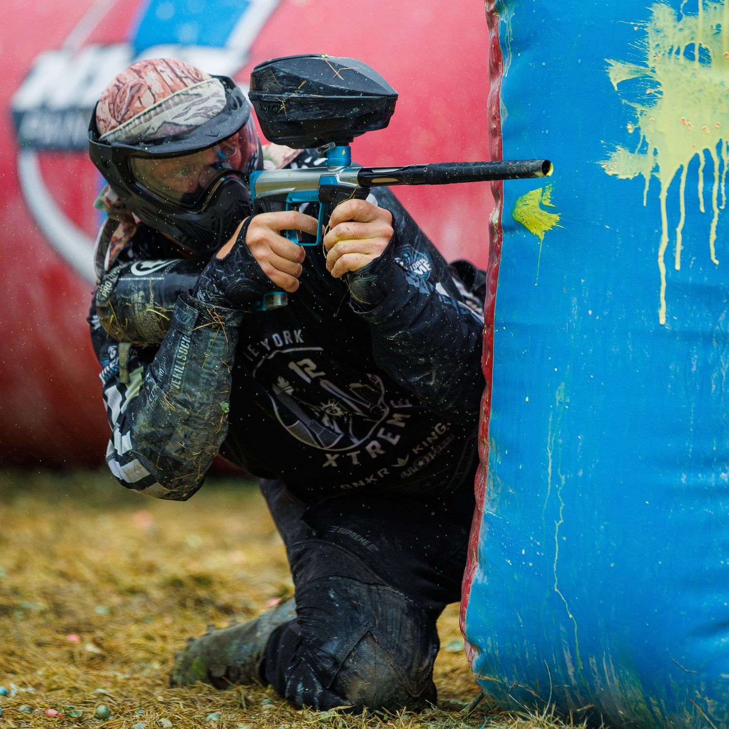 Paintball competitor in protective mask takes aim from behind blue inflatable bunker.