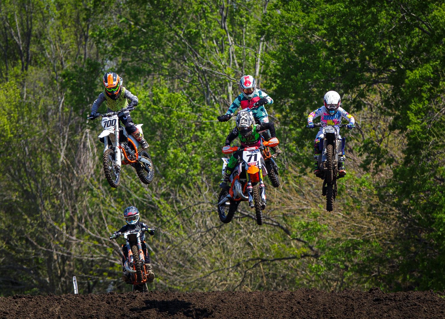 Four motocross riders soar through the air simultaneously on a wooded dirt track during a race.