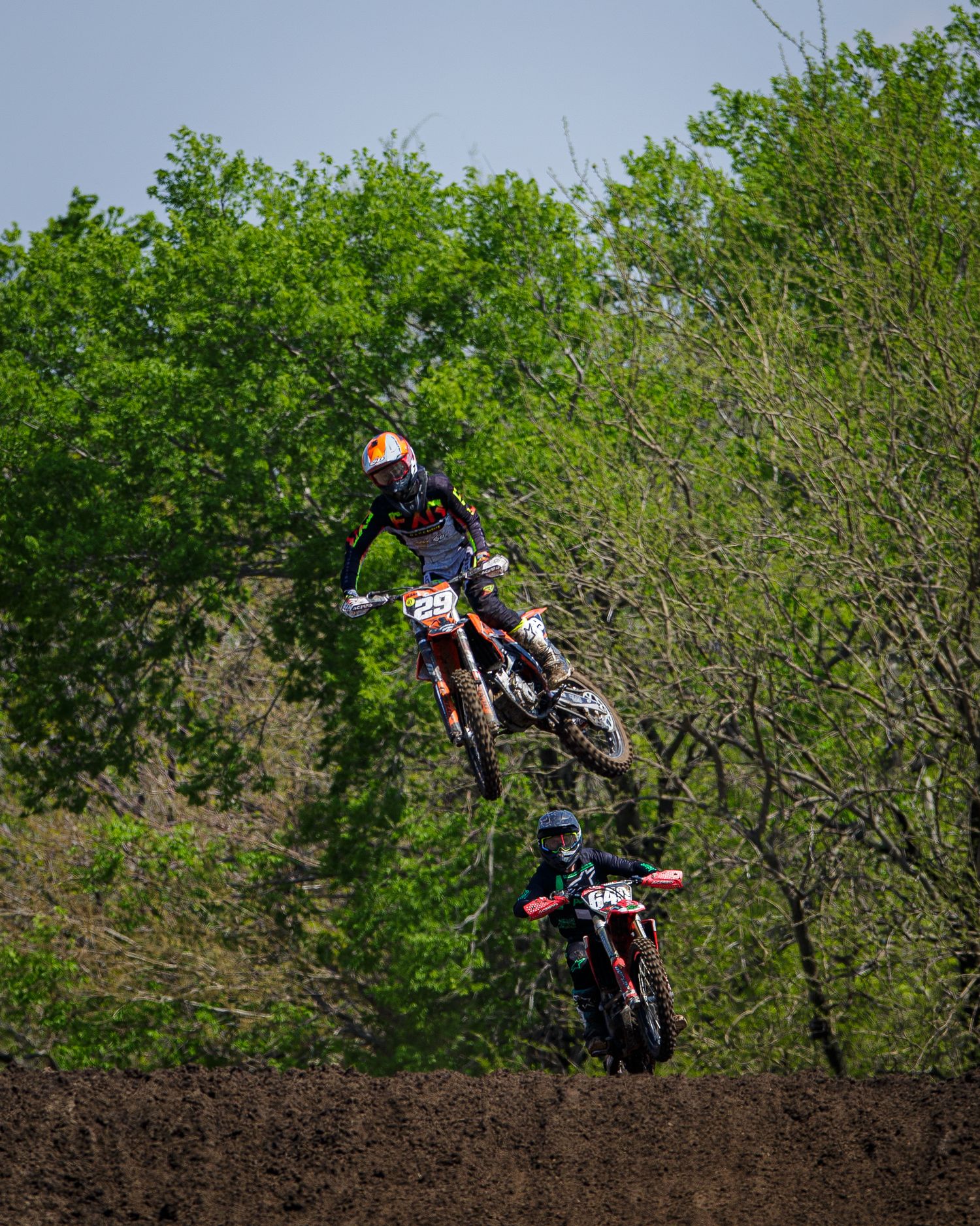 Three motocross riders execute sequential jumps on a dirt track surrounded by green foliage.