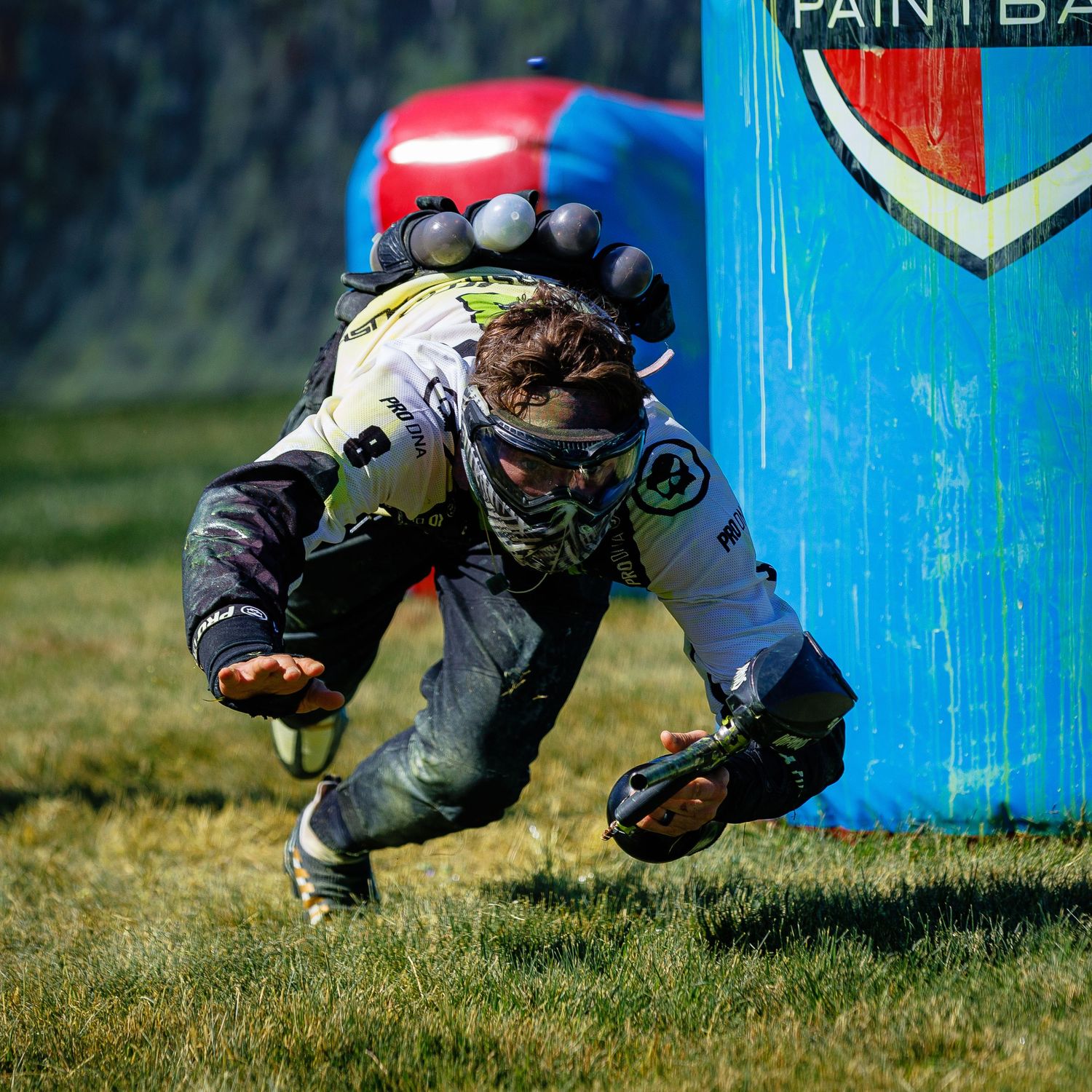 Paintball player diving behind a blue bunker during competitive speedball match.