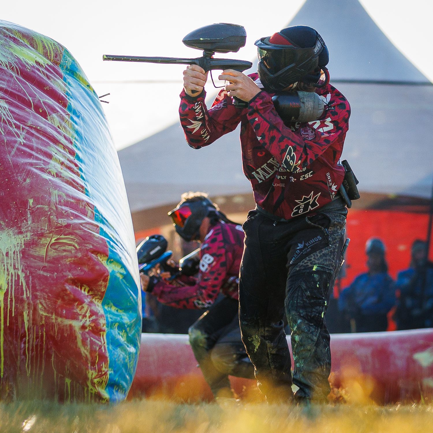 Paintball player in red jersey taking aim behind an inflatable bunker during a match.