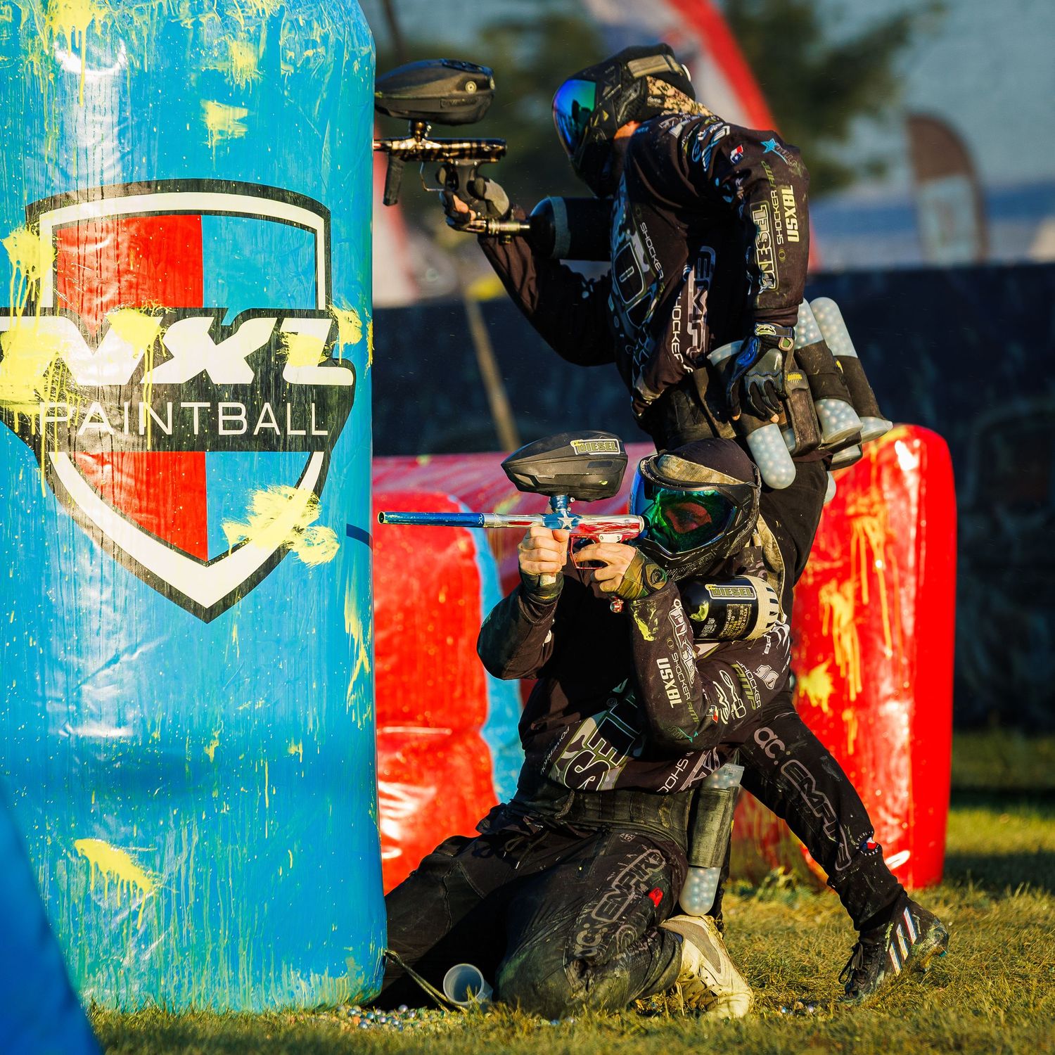 Paintball players in tactical positions shoot from behind inflatable field bunkers.