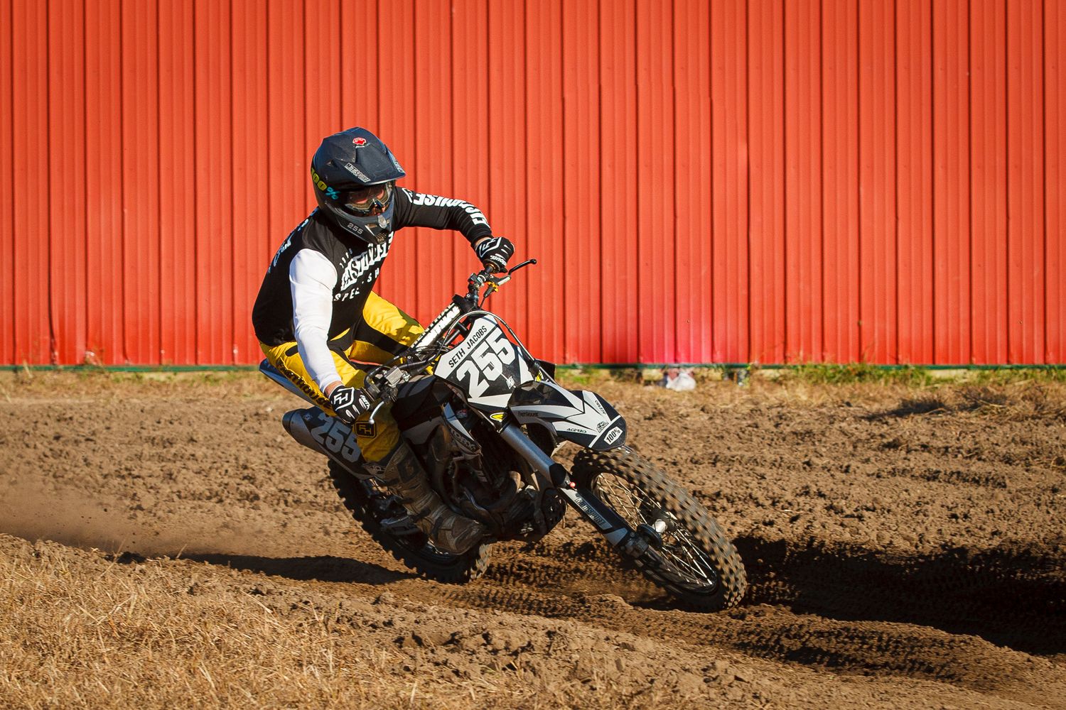 Dirt bike rider leans into turn against orange metal wall background.