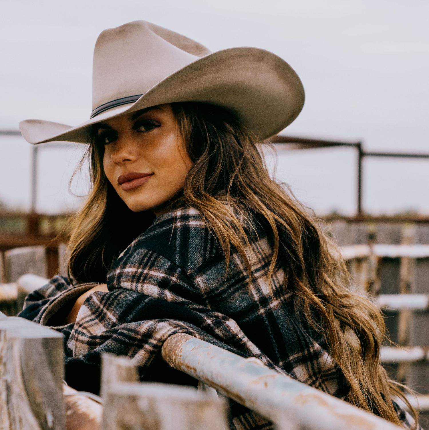 Woman in western attire with a cowboy hat leaning on a fence at sunset.