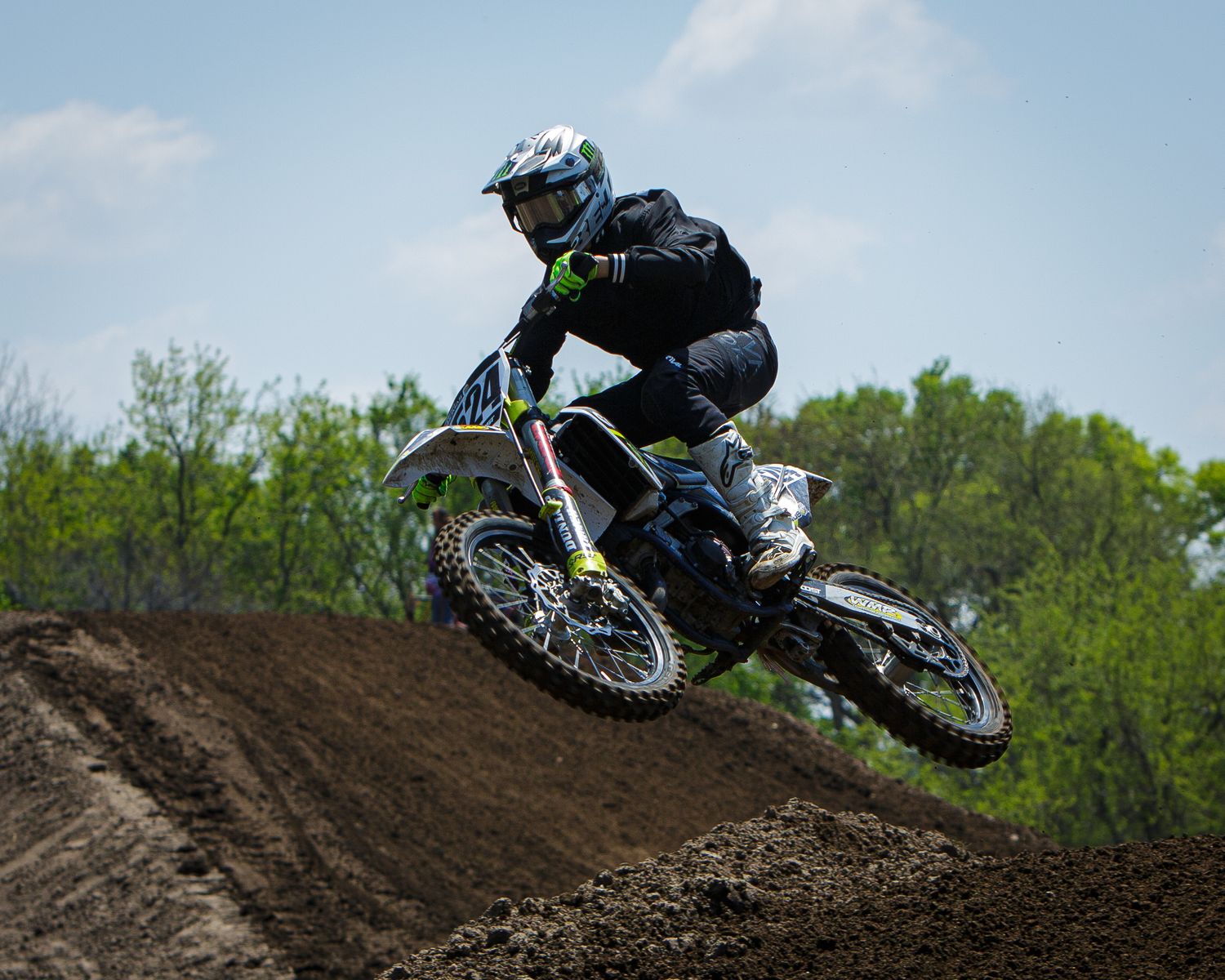 Dirt bike rider launches off muddy jump during motocross race through wooded course.