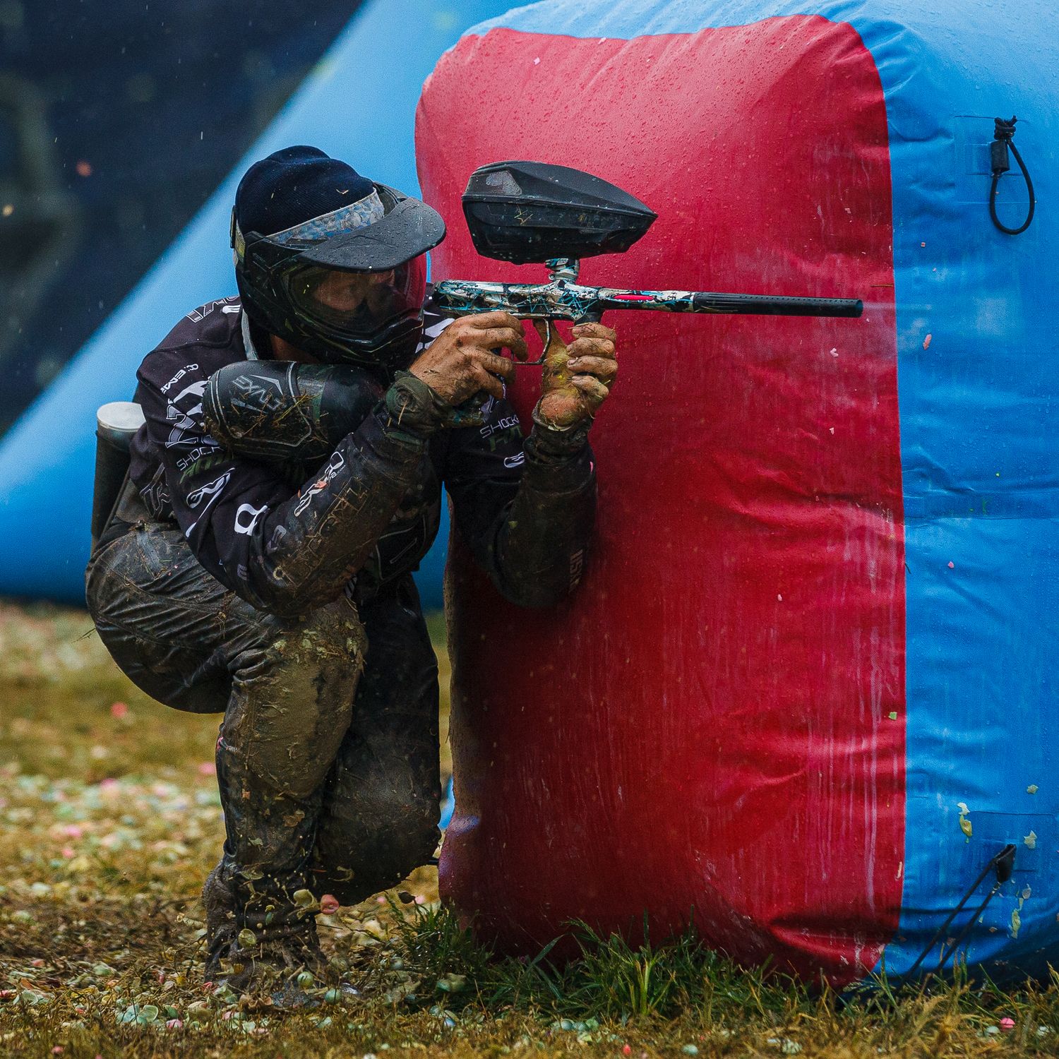 Paintball player in dark gear takes cover behind red inflatable bunker during game.