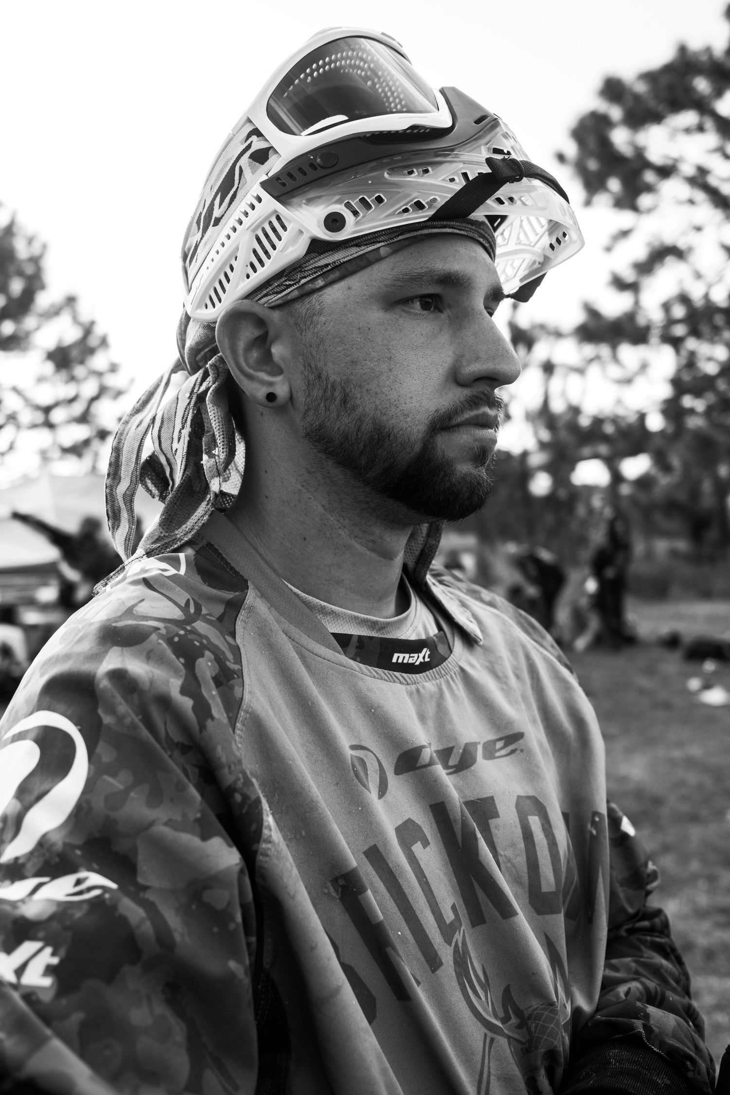 Paintball player in protective gear and camo jersey looking to the side during outdoor game.