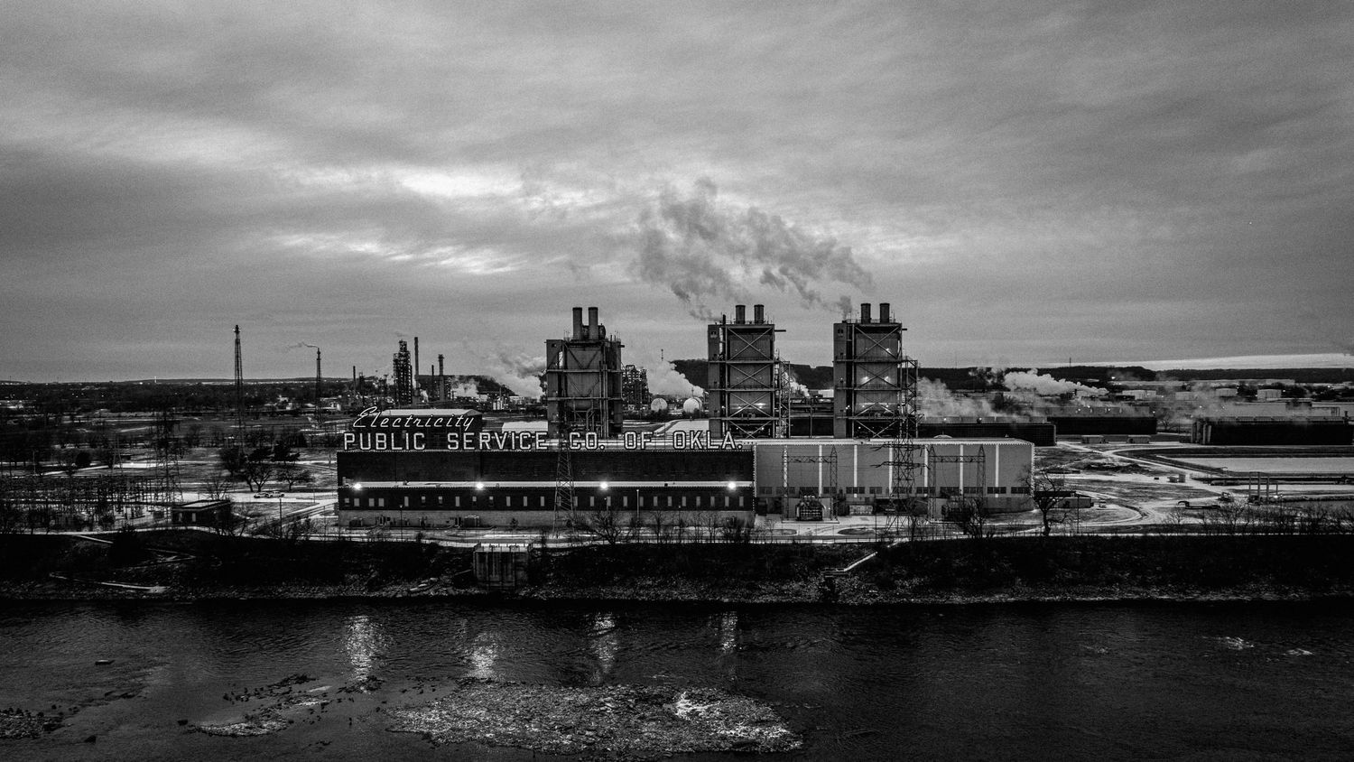Large industrial plant with smokestacks reflected in water under an overcast sky.