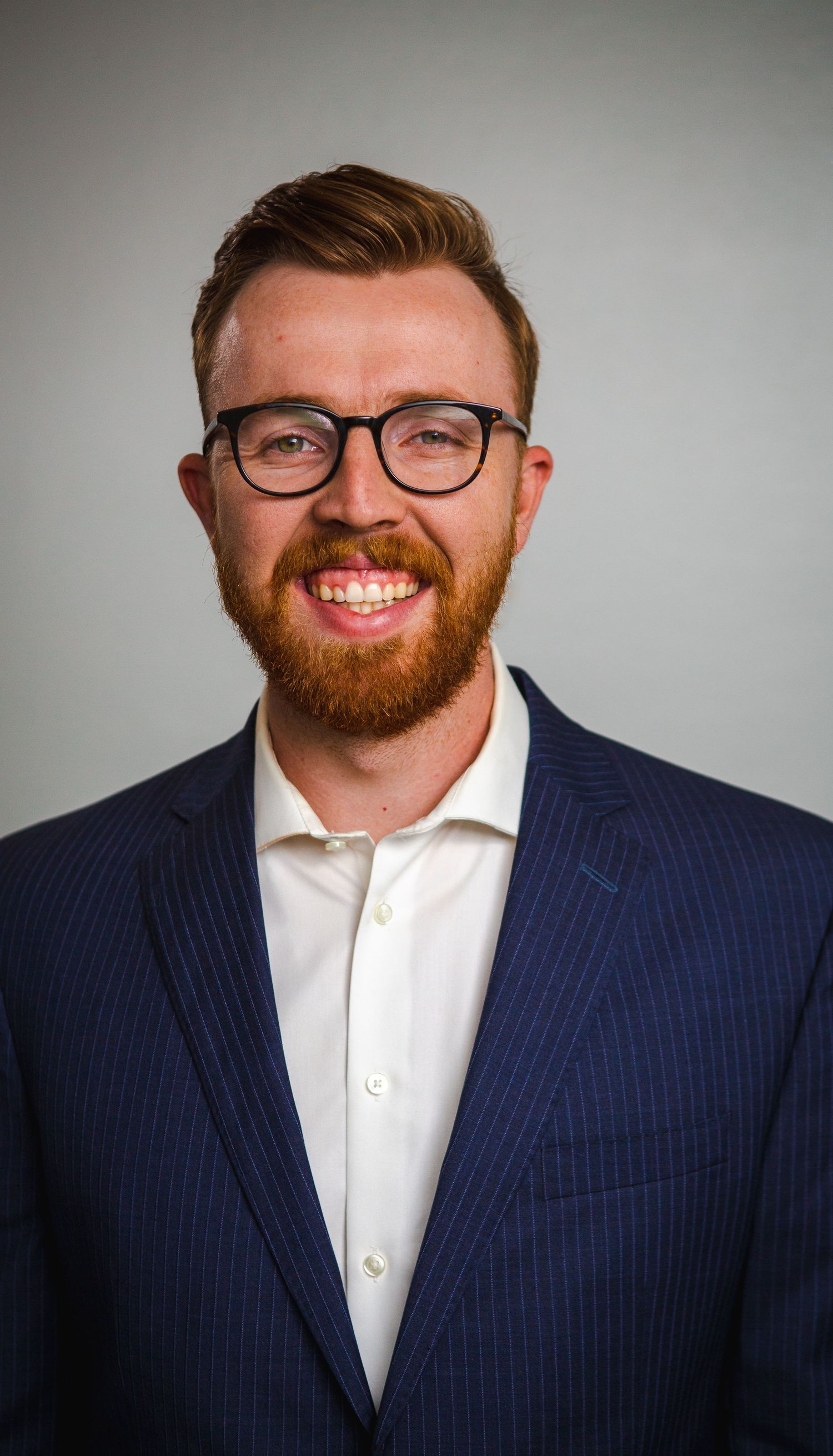 Professional headshot of person in navy suit and glasses against gray background.