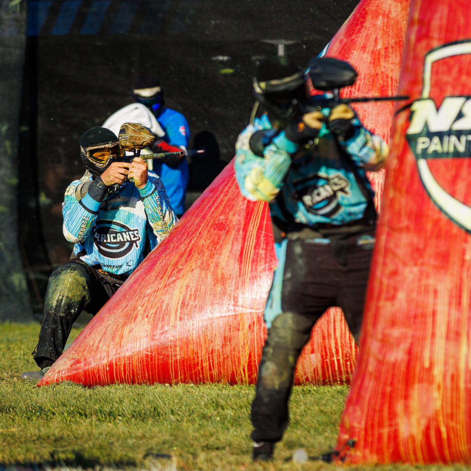 Paintball players engage in firefight behind red and blue inflatable bunkers during match.