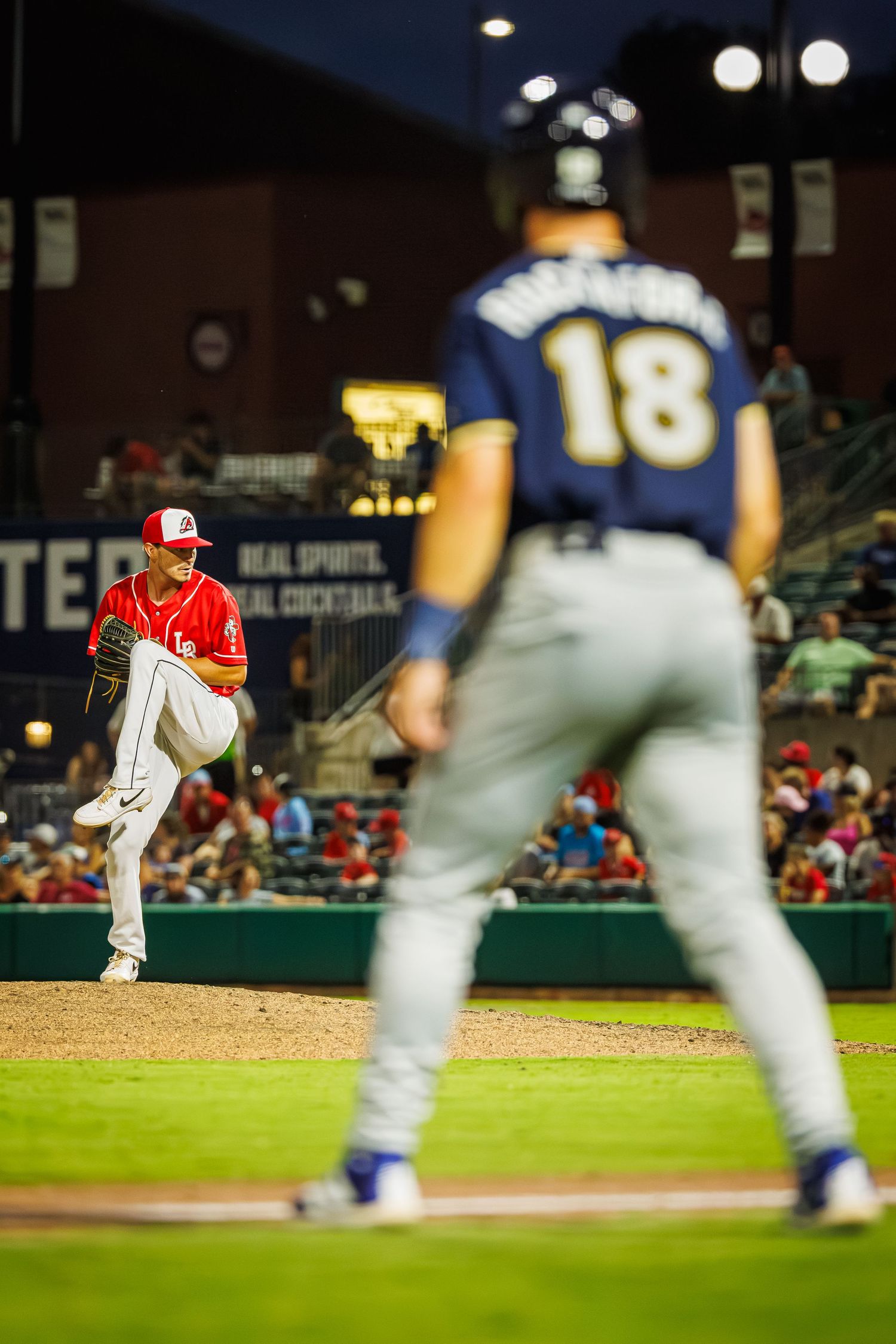 Baseball pitcher in red uniform delivers pitch from mound during night game with blurred players in foreground.