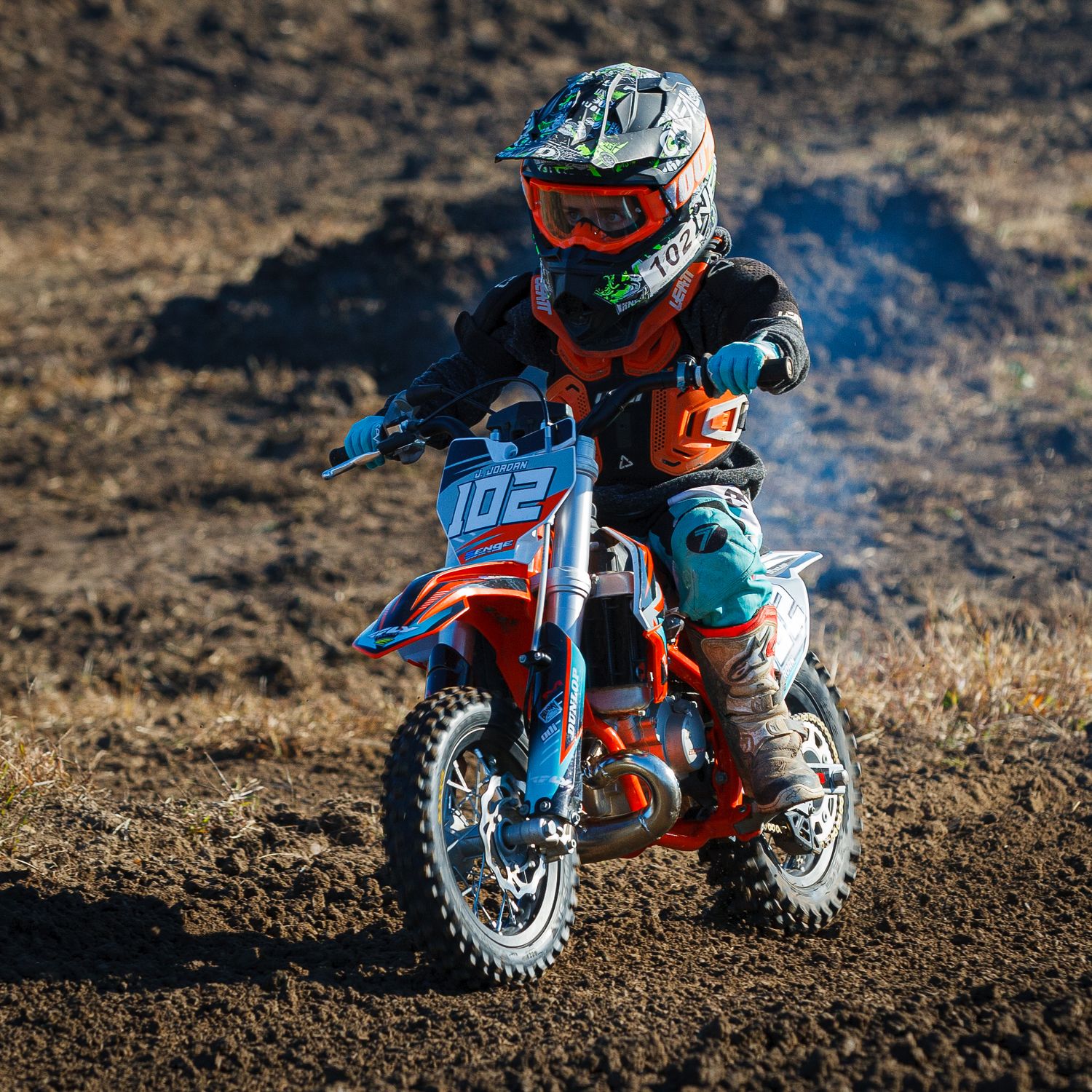 Young rider on small KTM dirt bike performs stunts on dusty track.