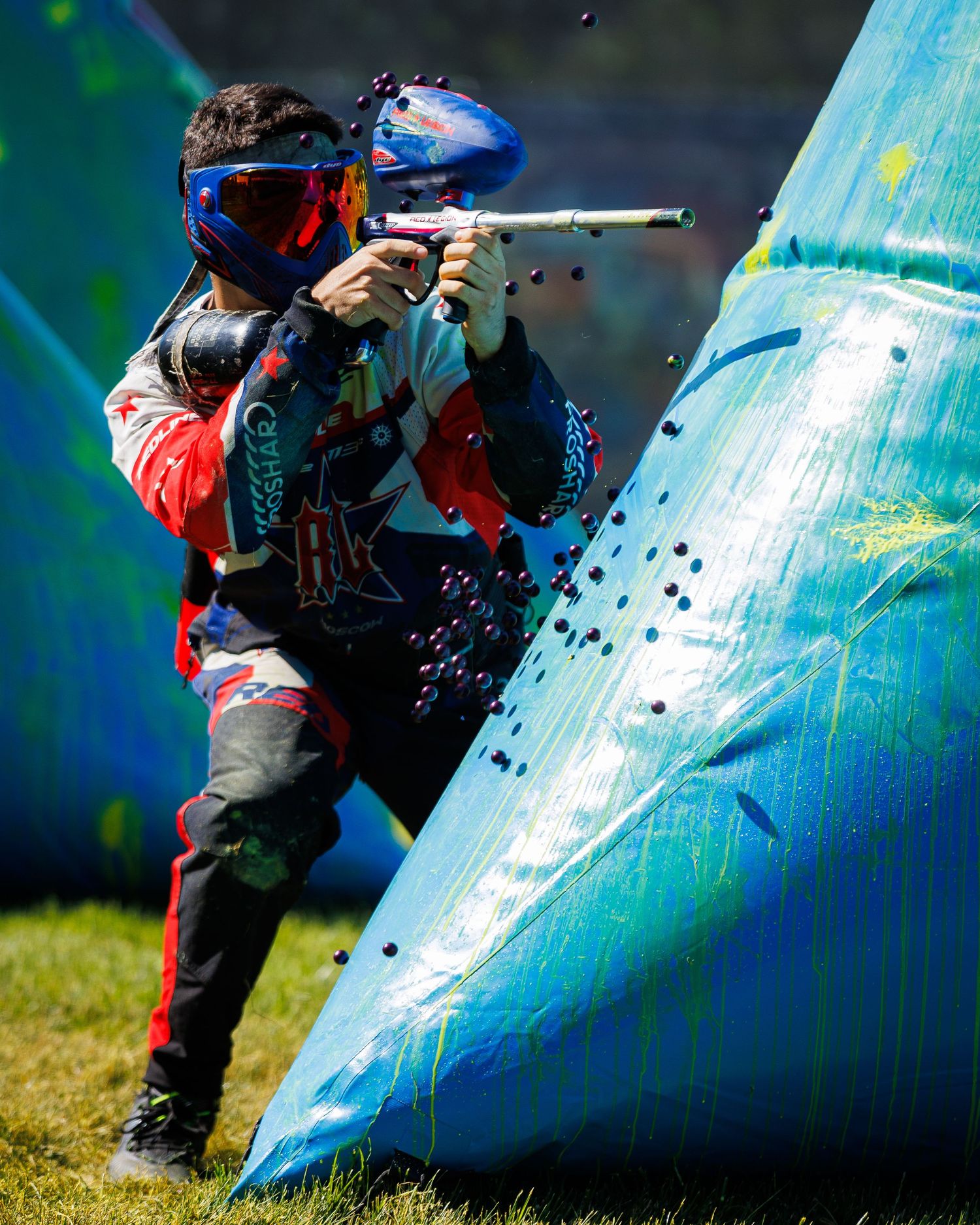 Paintball player in protective gear taking aim from behind a blue bunker during a match.