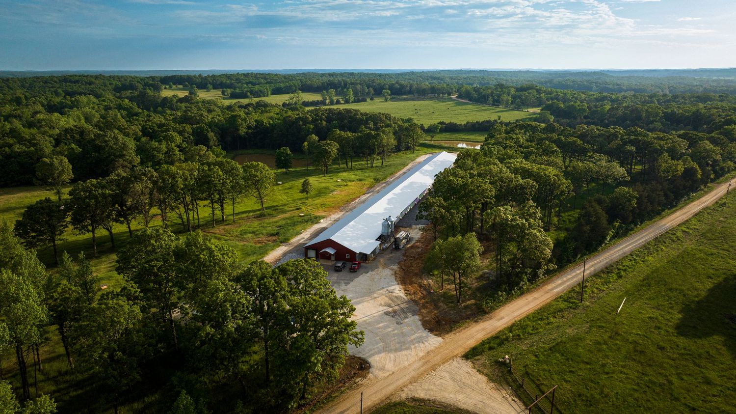 Long white agricultural building surrounded by green forest from aerial perspective.