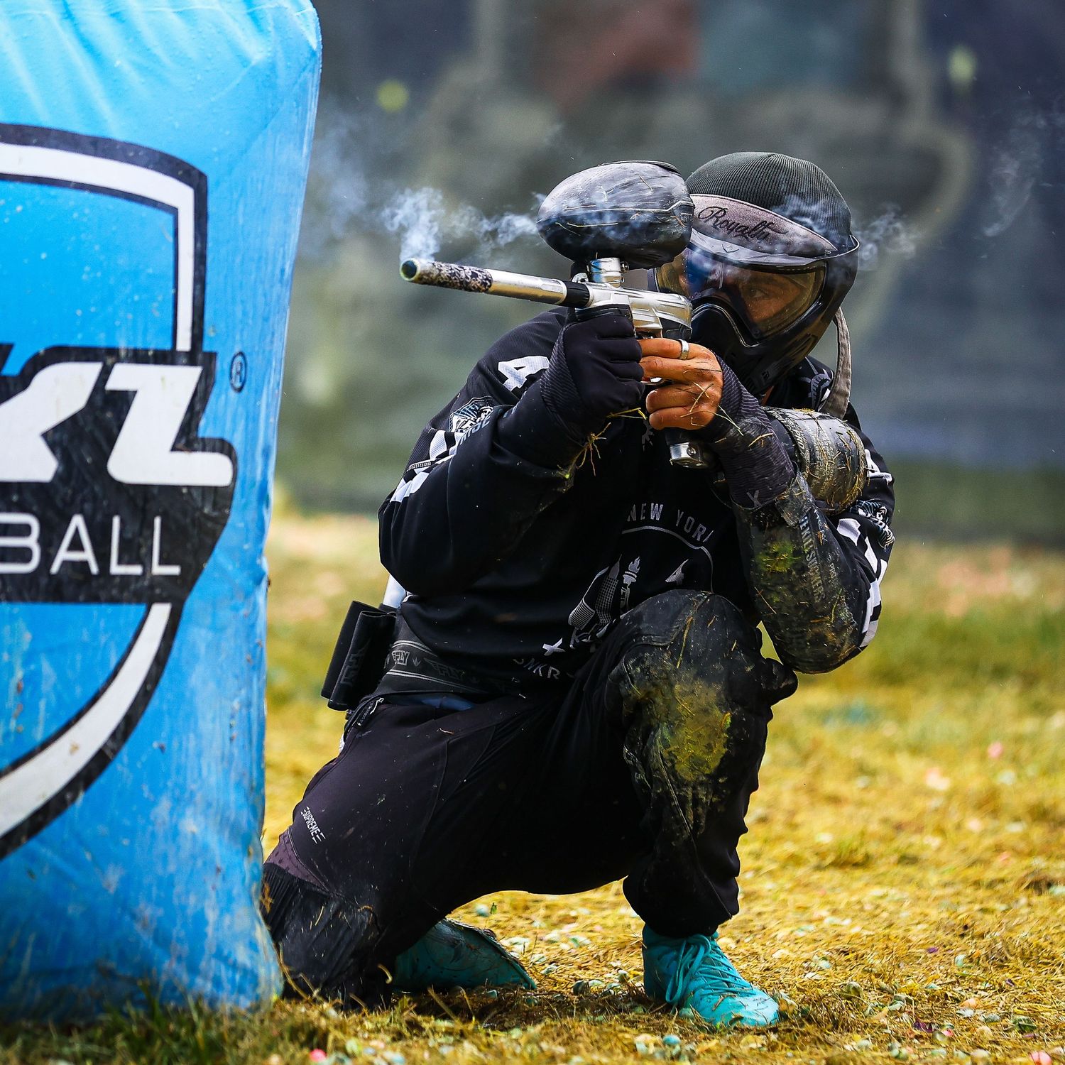 Paintball player in black gear crouches behind blue bunker while smoke rises from hits.