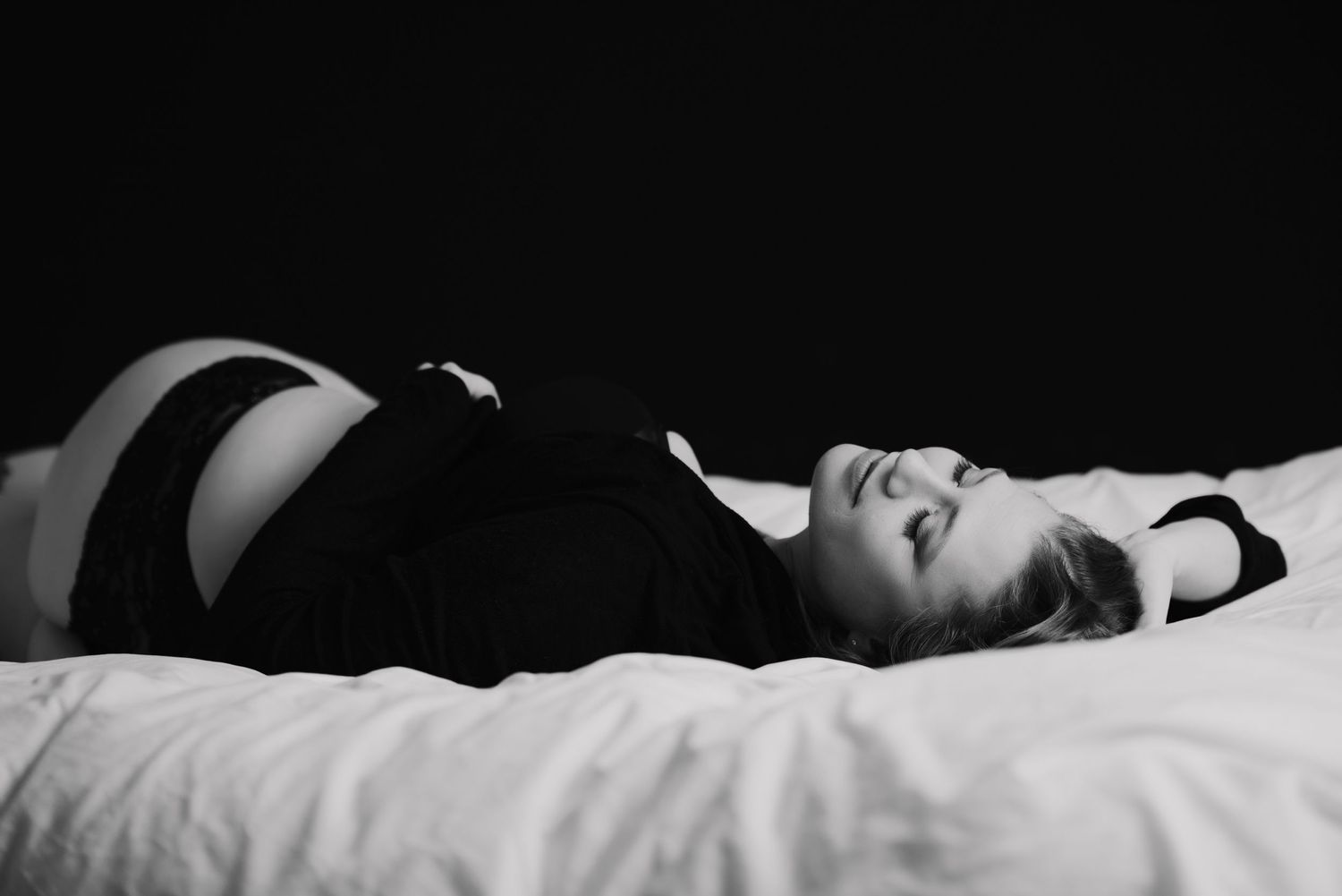 A woman in black lingerie lies gracefully on white bedding in a dramatic black and white photograph.