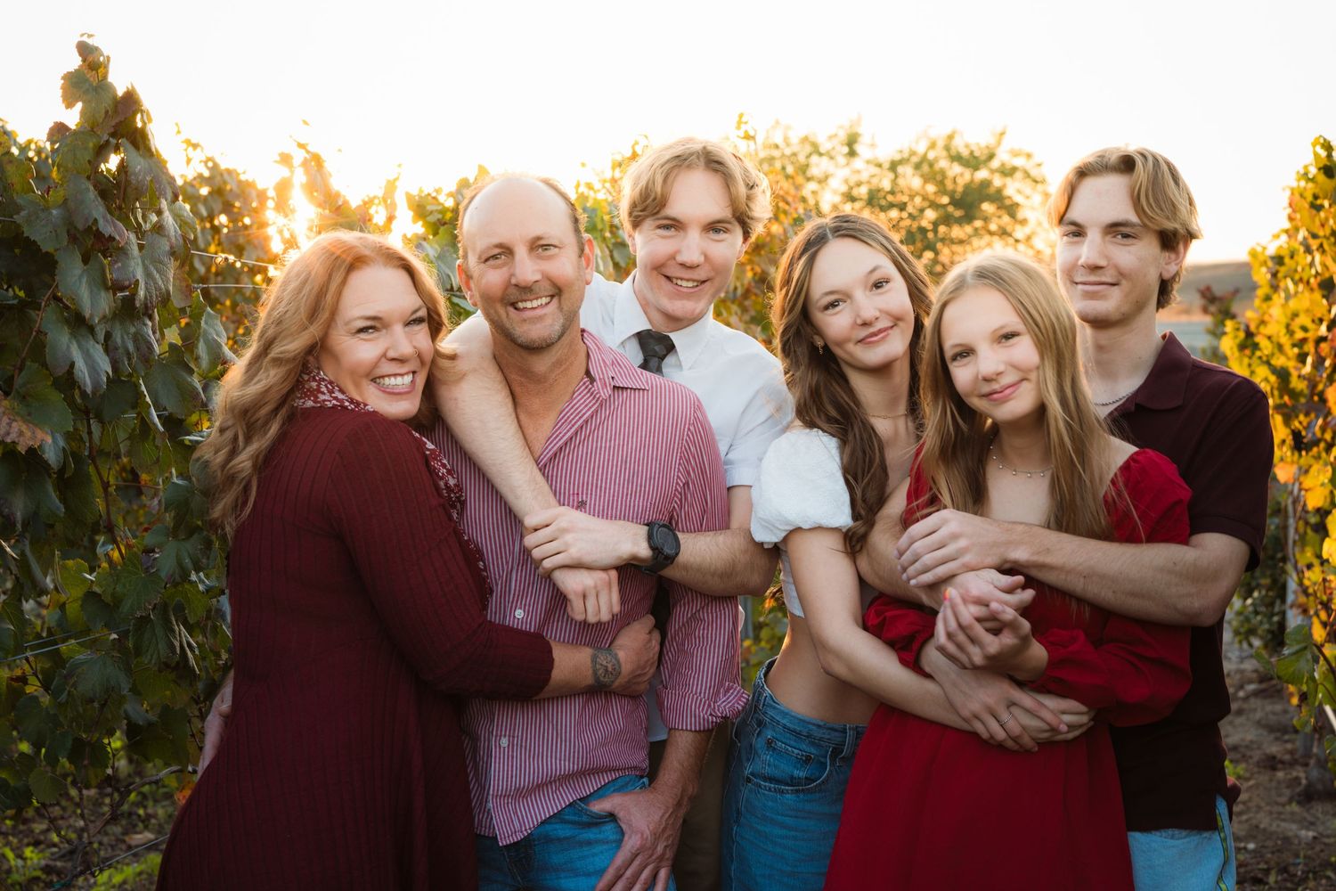 Happy family of six posing together in a vineyard during golden hour, wearing coordinated burgundy and pink outfits.