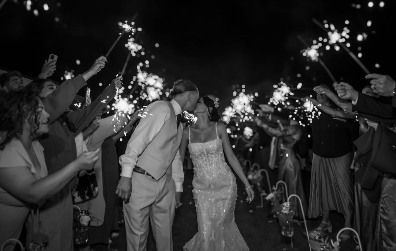 Wedding couple exits through sparkler tunnel held by guests during nighttime celebration in black and white photograph.