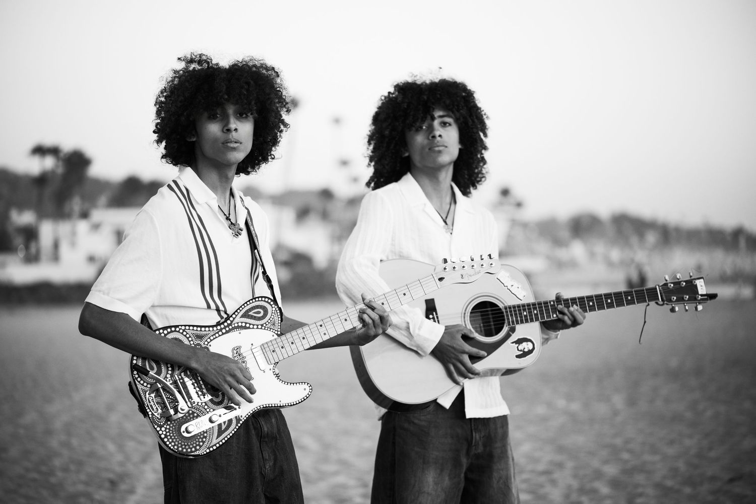 Two musicians with curly hair playing electric and acoustic guitars on a beach in black and white photo.