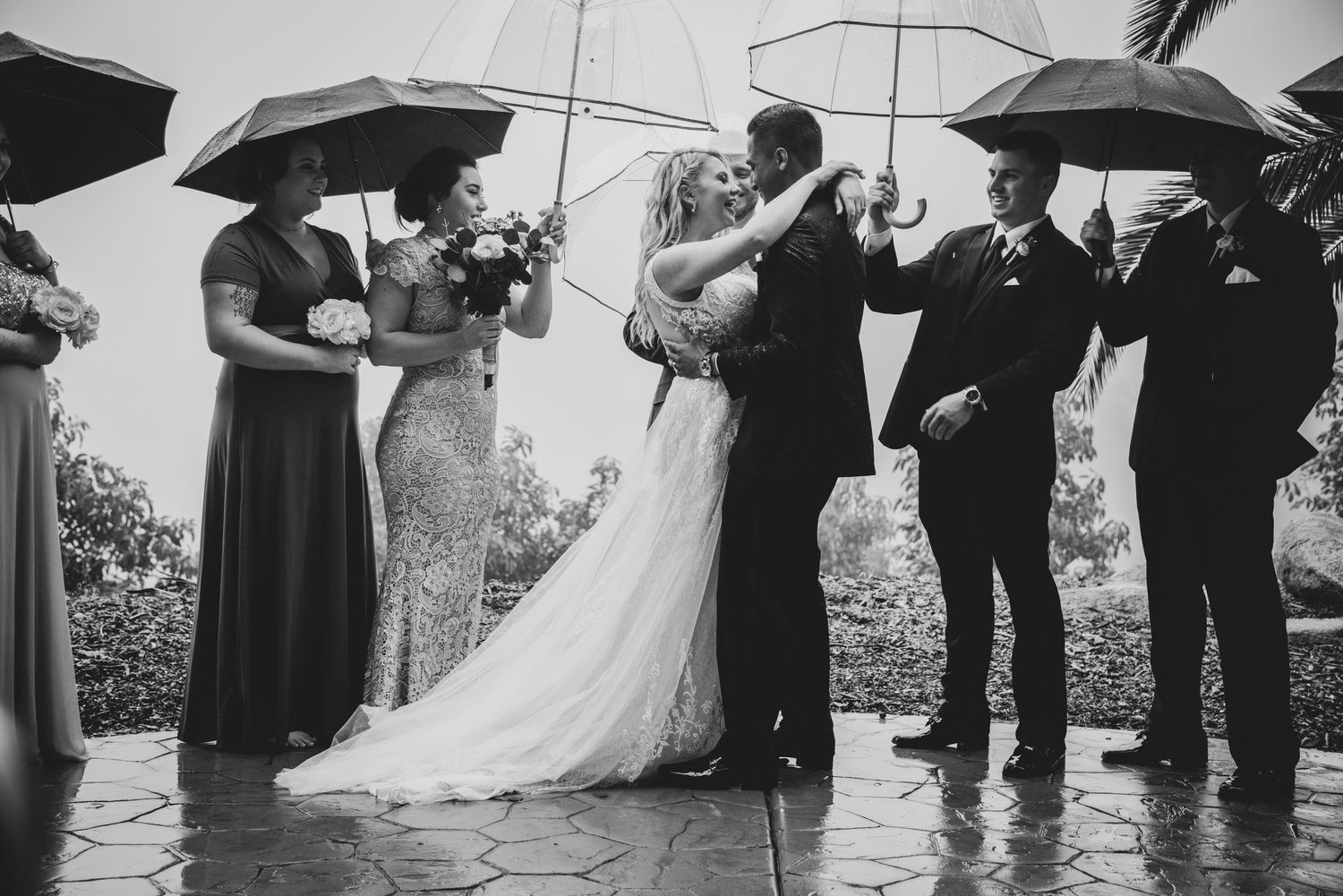 Black and white wedding photo showing a group with umbrellas during a rainy outdoor ceremony.